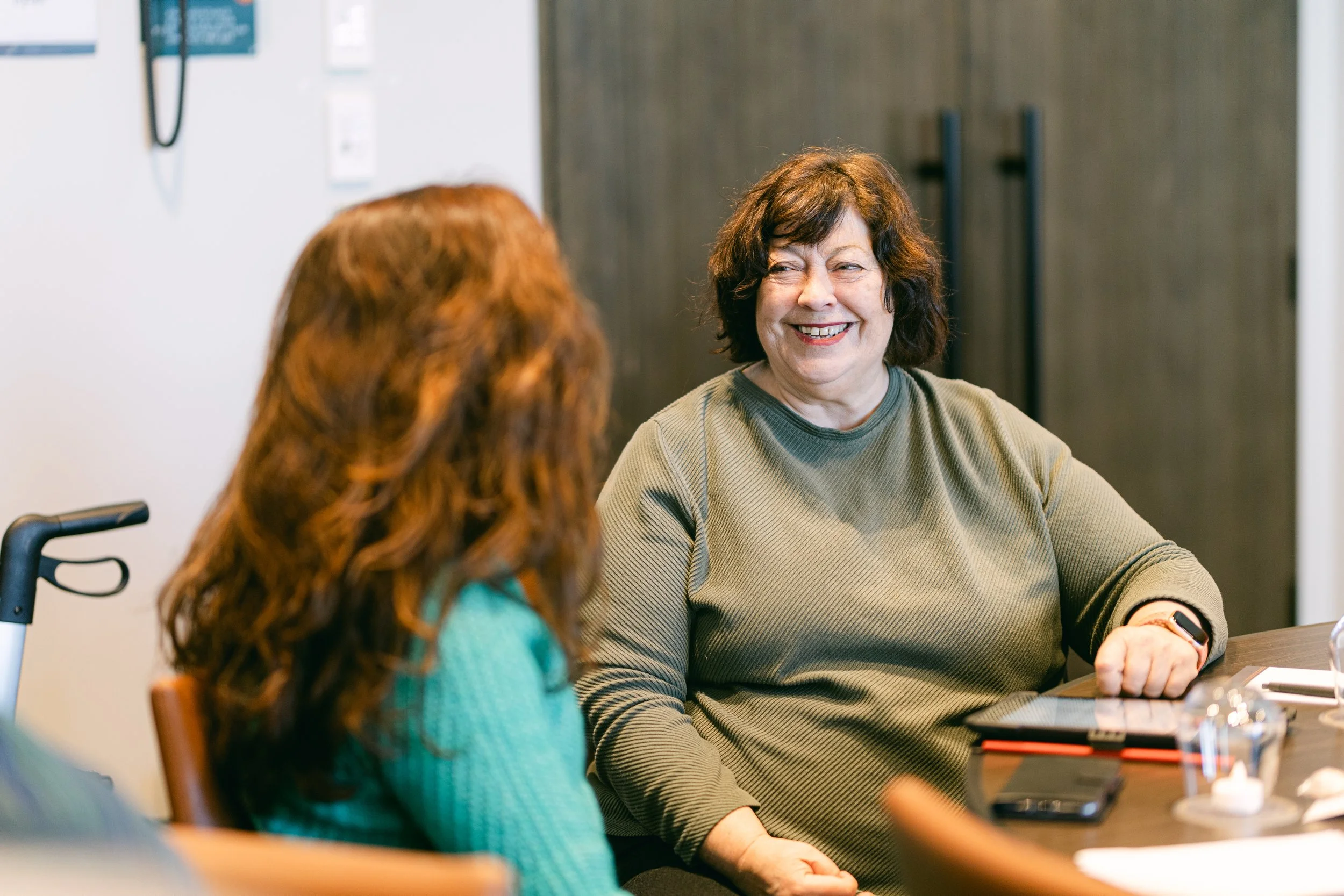 Two women sitting at a table, smiling and talking, in an indoor setting.