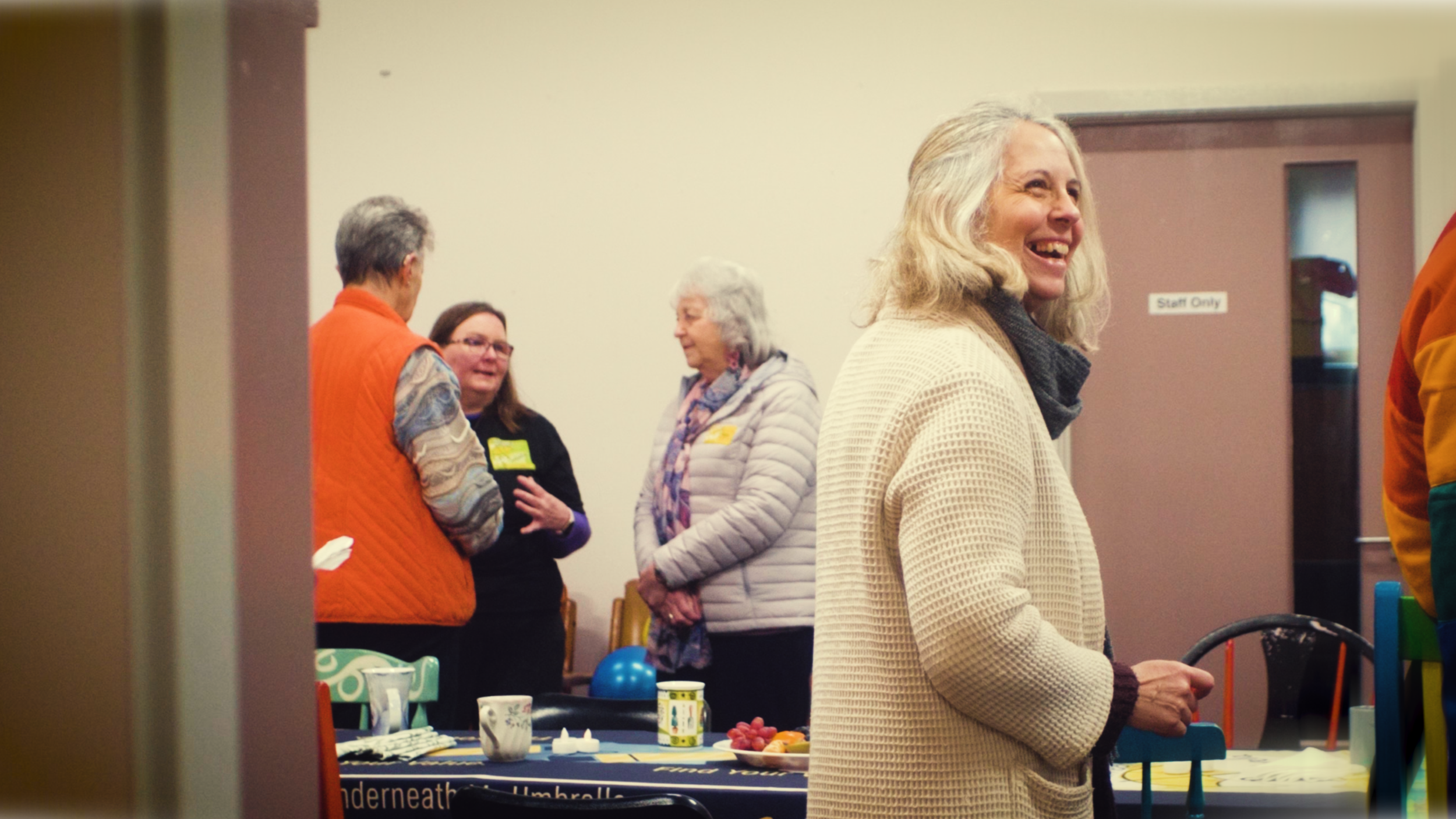 A group of four women and one partially visible person in a room, talking and smiling, with a table of snacks and cups in front of them. One woman in the foreground is wearing a cream-colored sweater and has gray hair. The background features a door 