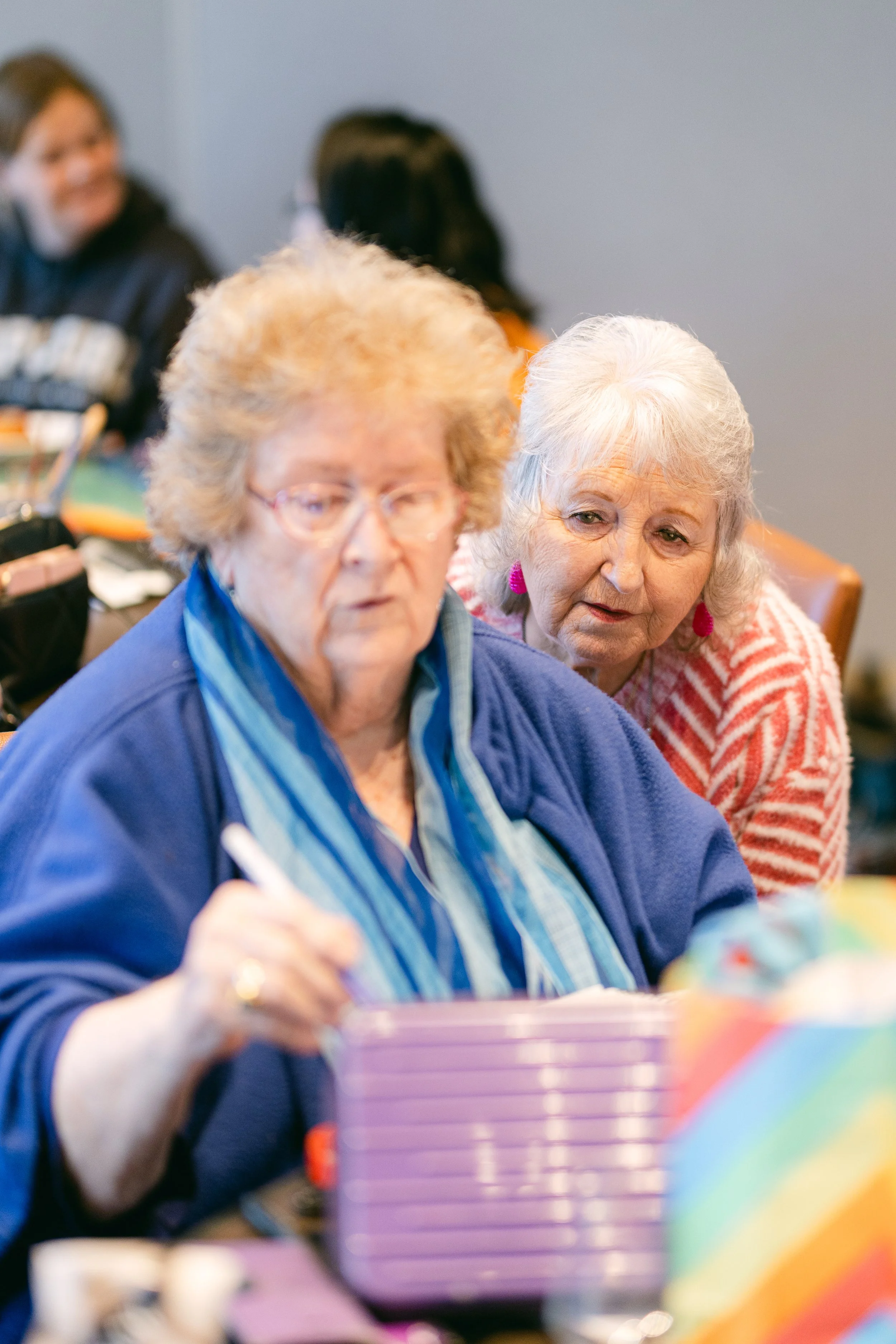Two elderly women seated at a table, one is writing or signing and the other is leaning in to look, with several colorful objects and books on the table.