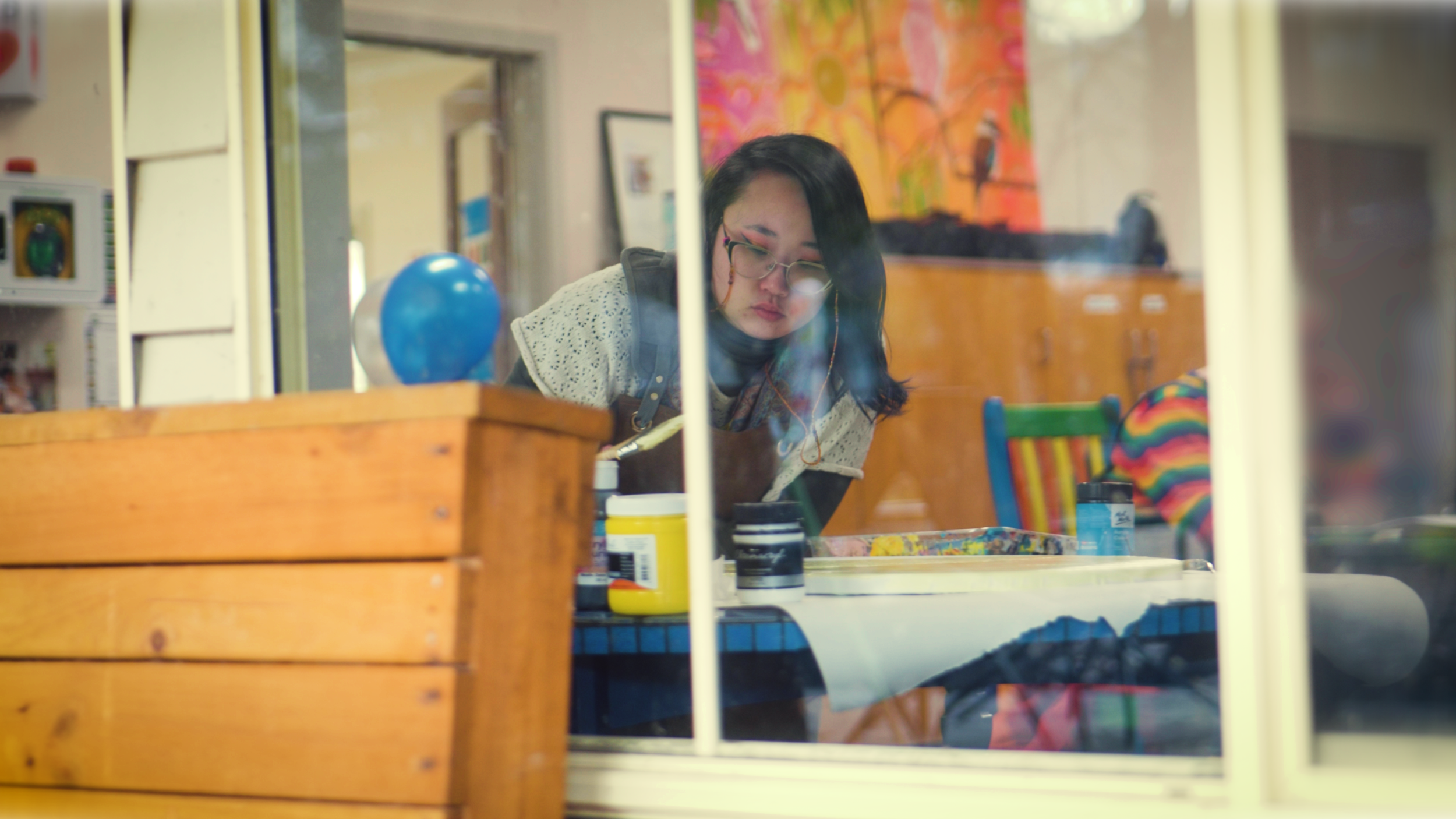A woman with glasses and dark hair, wearing a beige crochet sweater and an apron, is seen through a window, working on an art project at a table with paint supplies in a colorful, decorated room.