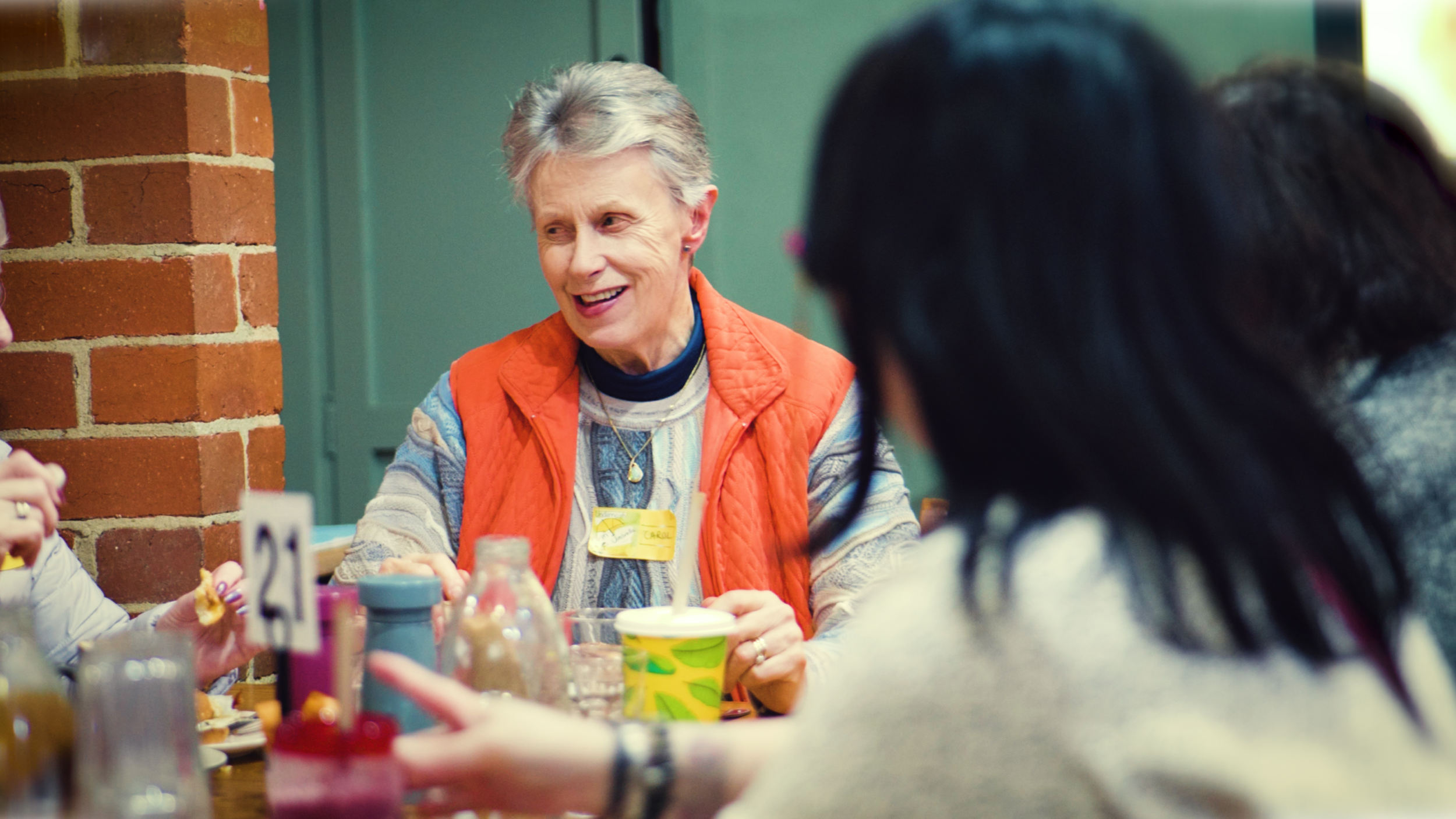 A smiling elderly woman with short gray hair talking to a group at a table in a restaurant or cafe. She is wearing an orange vest over a patterned shirt and a necklace with a small pendant.