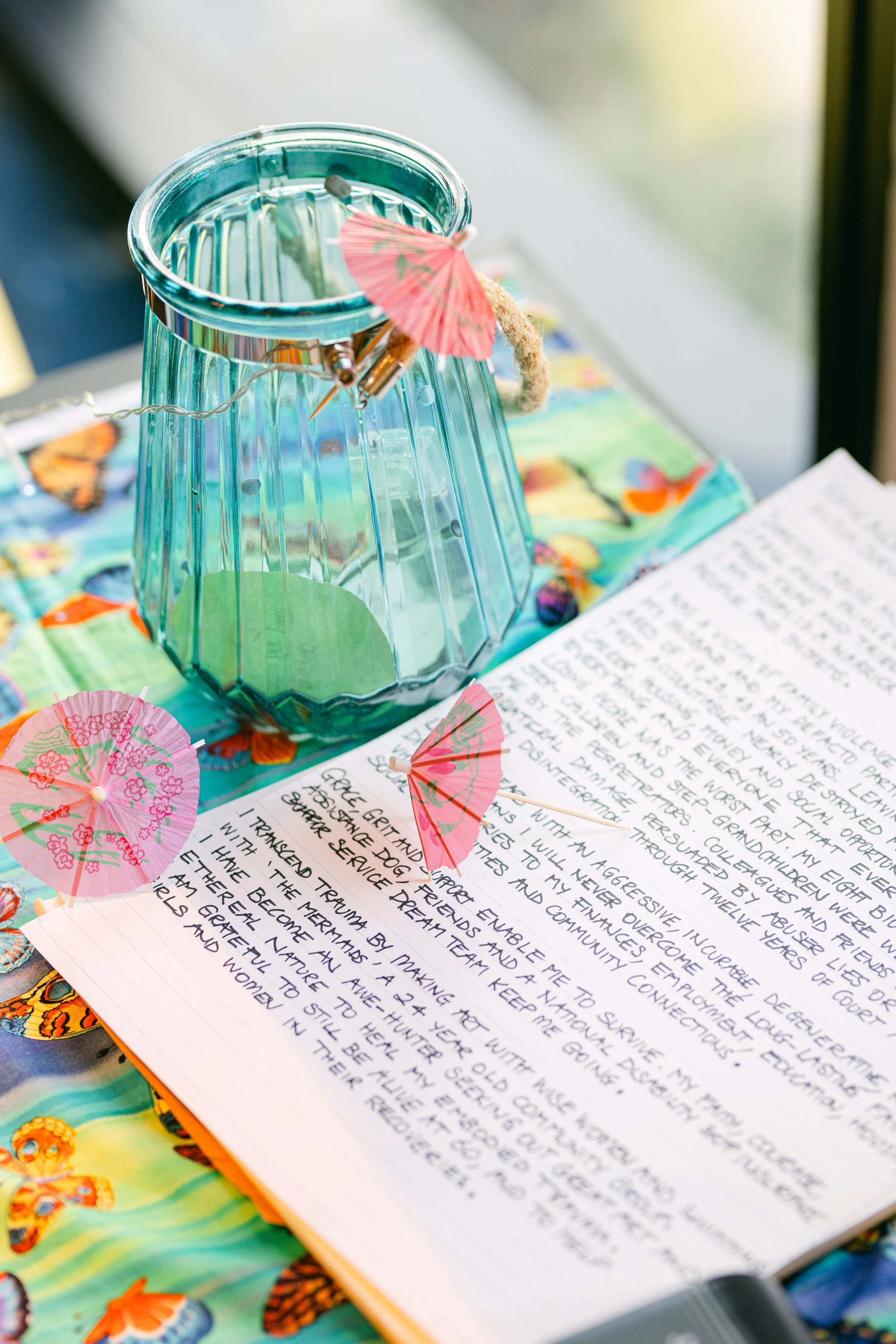 A glass lantern decorated with small pink paper umbrellas, placed on colorful butterfly-printed fabric. There is a handwritten journal or notebook underneath with visible text about personal reflections, near the lantern.