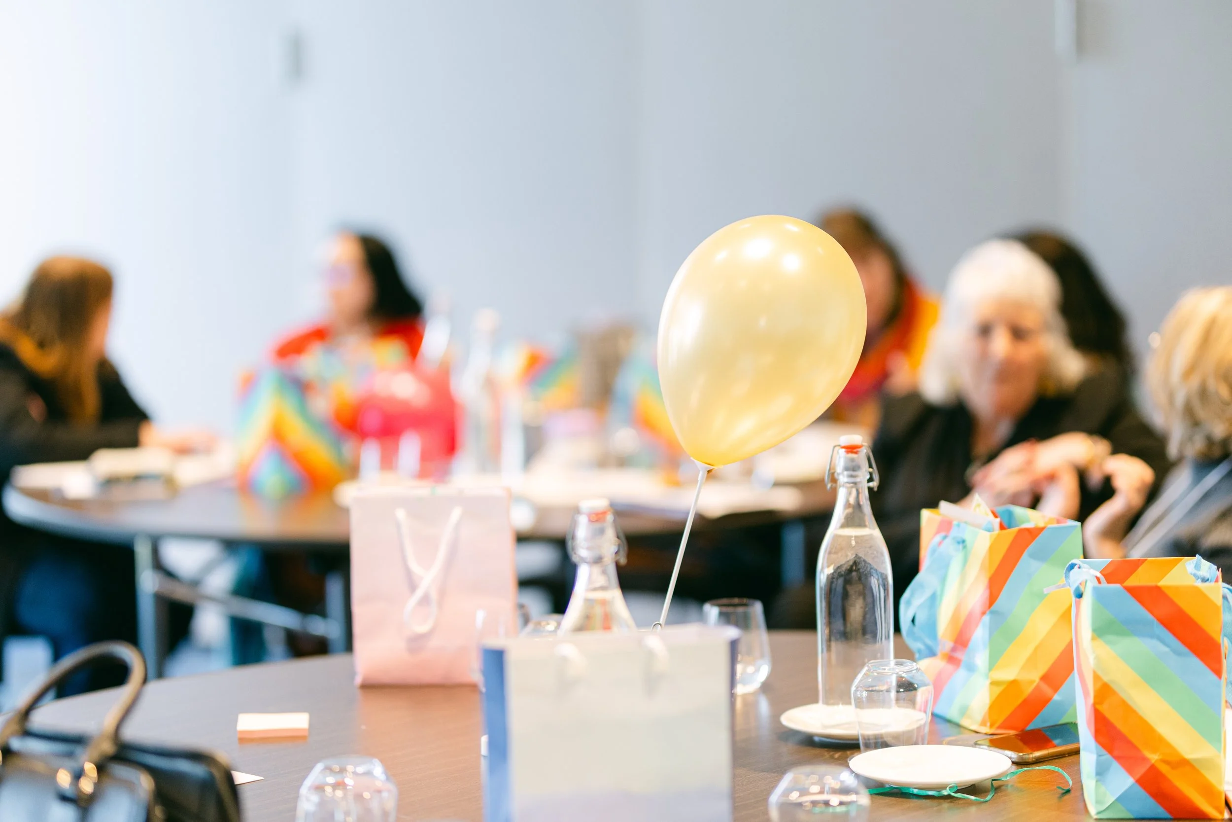 Party table decorated with rainbow-colored gift bags and a gold balloon in the center, with people in colorful attire in the background.