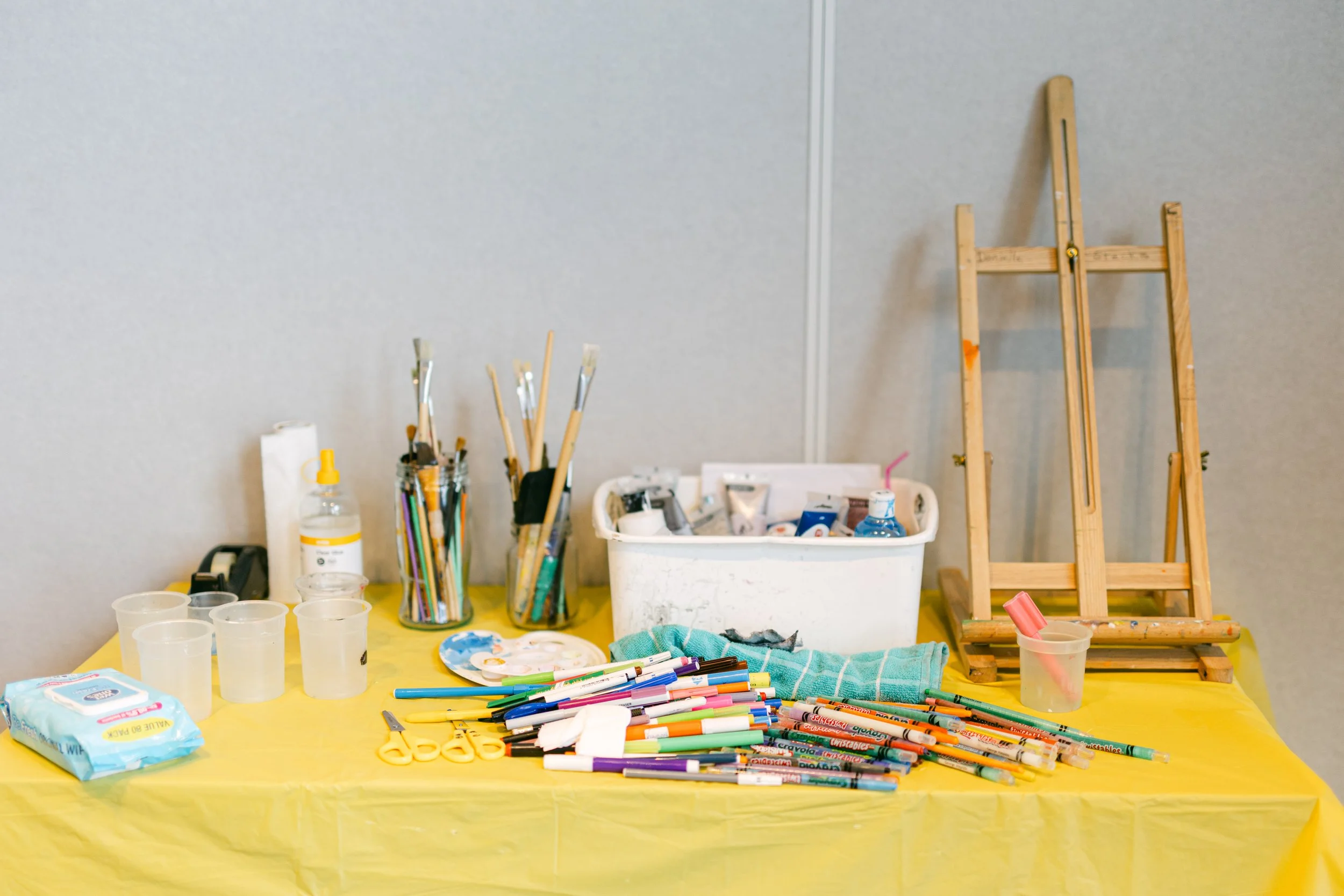 An art supplies table with various paintbrushes, markers, scissors, and other tools, a white container filled with art materials, a small easel, and snacks on a yellow tablecloth against a gray wall.
