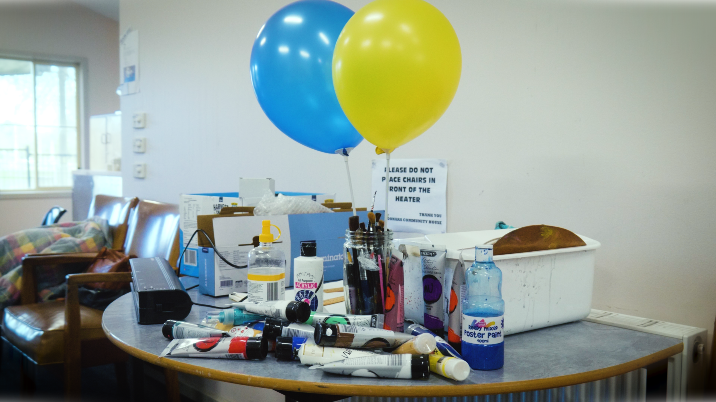 Table with paint supplies, including tubes of paint, brushes, a bottle of poster paint, and balloons, in a room with chairs and a window.