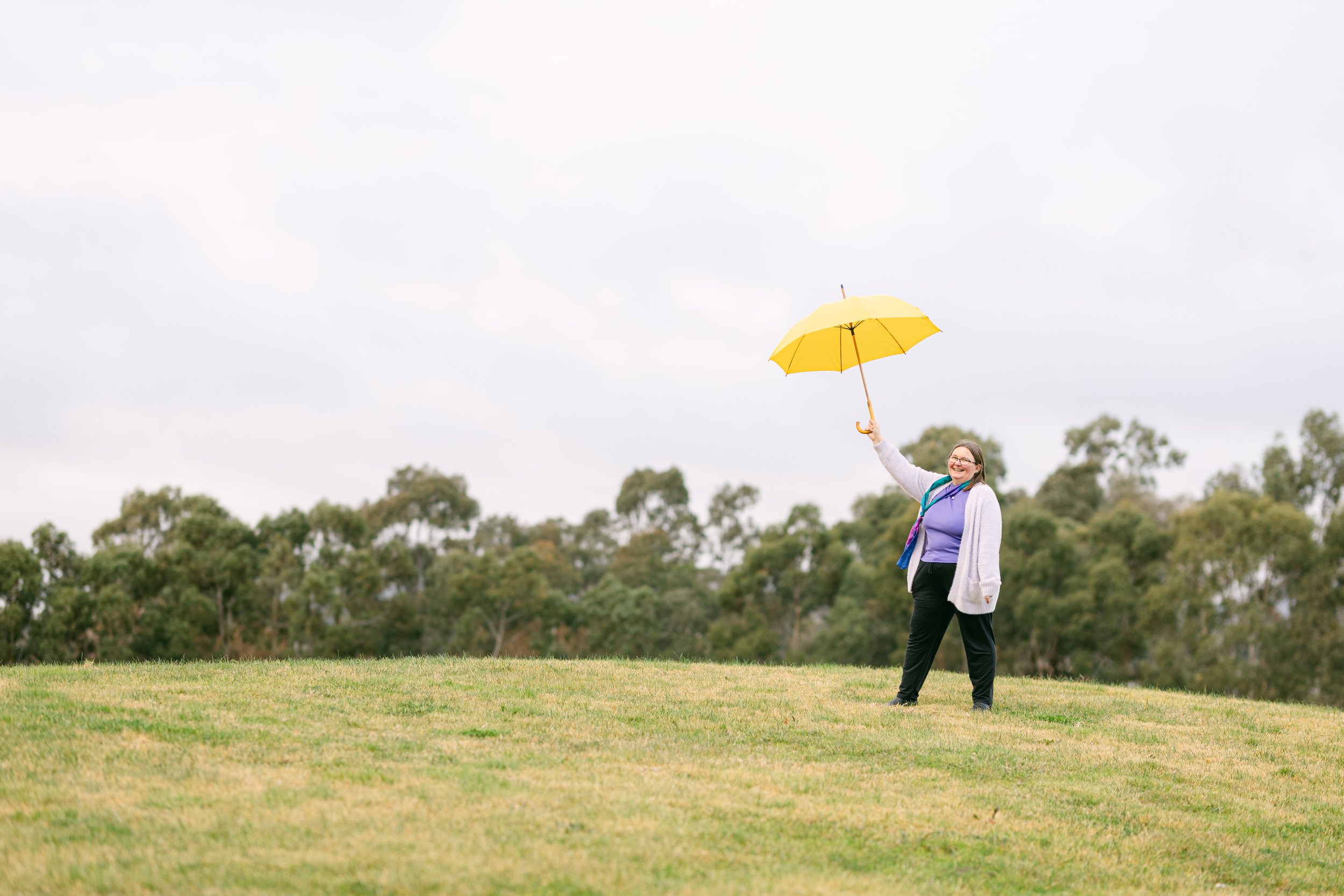 Person holding a yellow umbrella outdoors on a cloudy day, surrounded by green trees and grass.