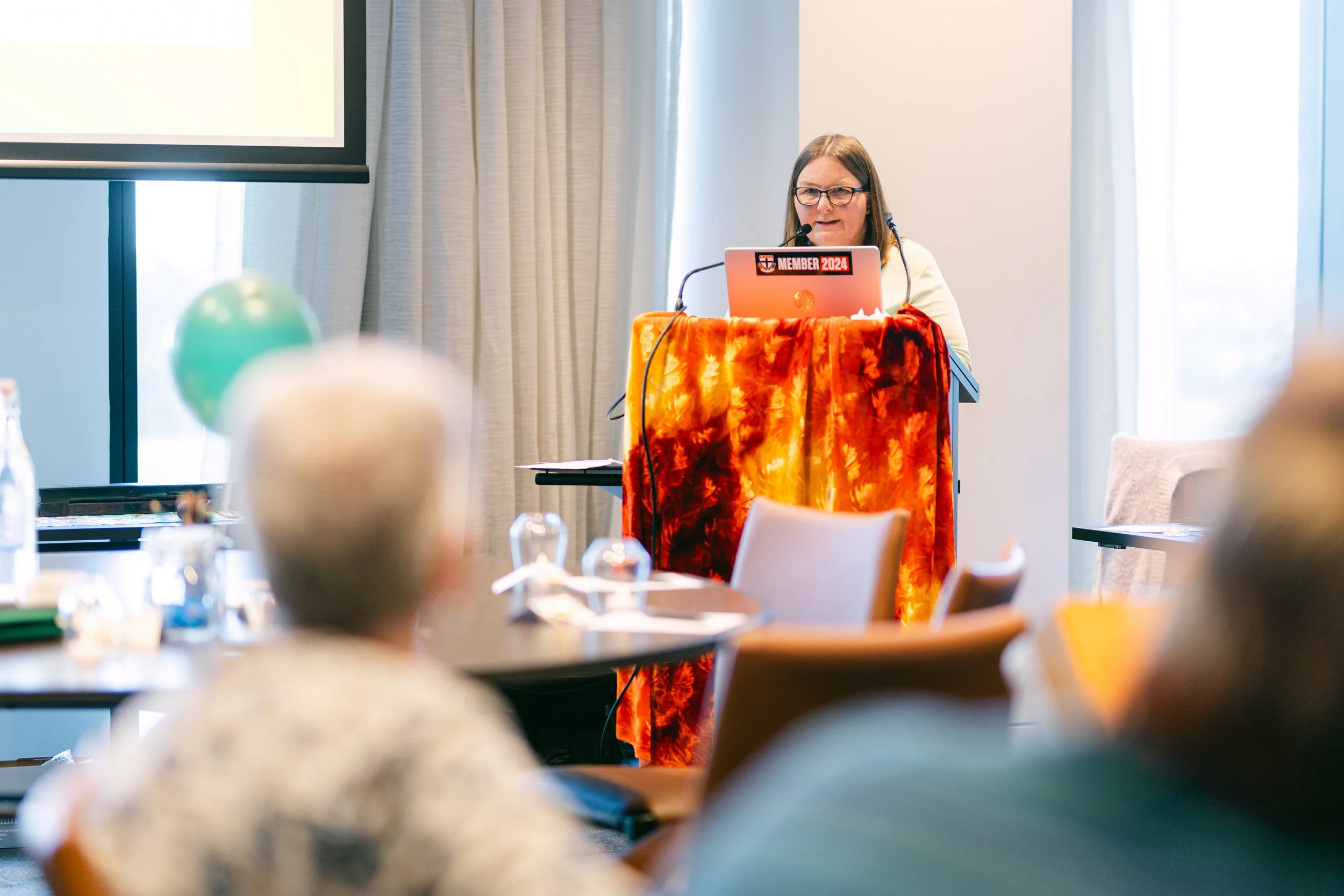 A woman standing at a podium giving a presentation in a conference room, with a laptop that has a sticker. The room has beige curtains, a window allowing natural light, and round tables with seated attendees in the foreground.