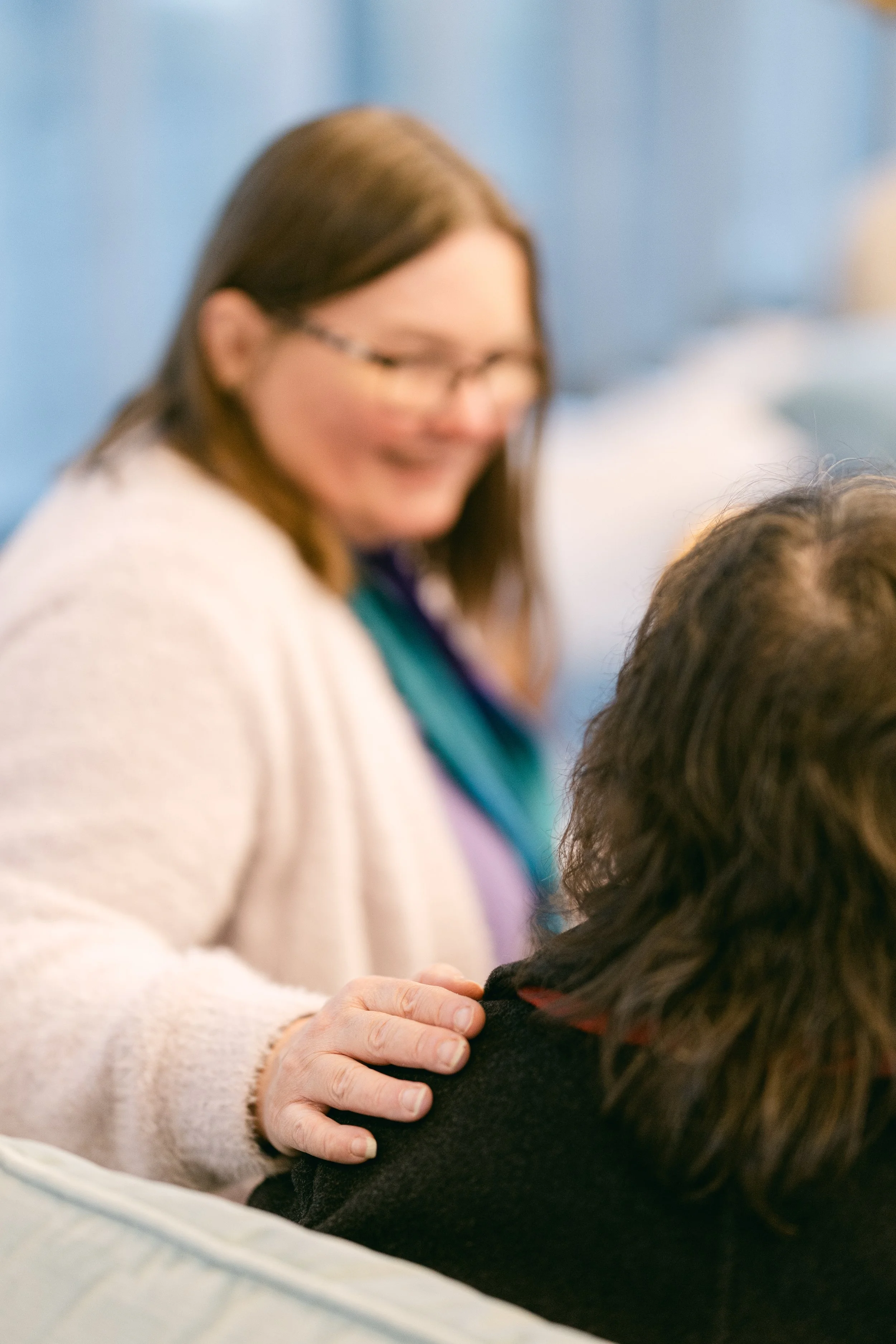 A woman with glasses gently places her hand on another person's shoulder, smiling warmly.