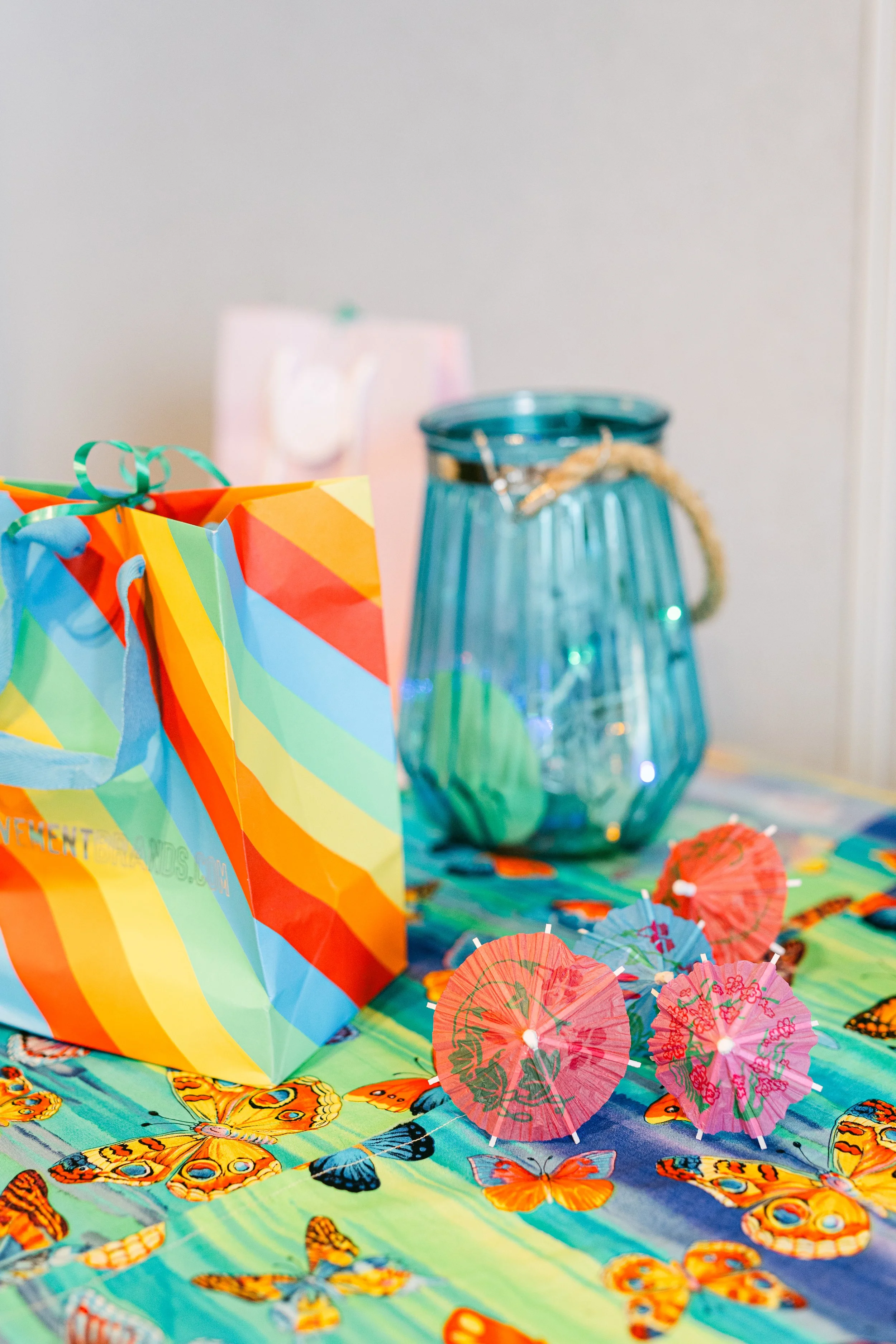 Colorful birthday table with butterfly-themed tablecloth, paper umbrellas, a large glass container, and gift bags.