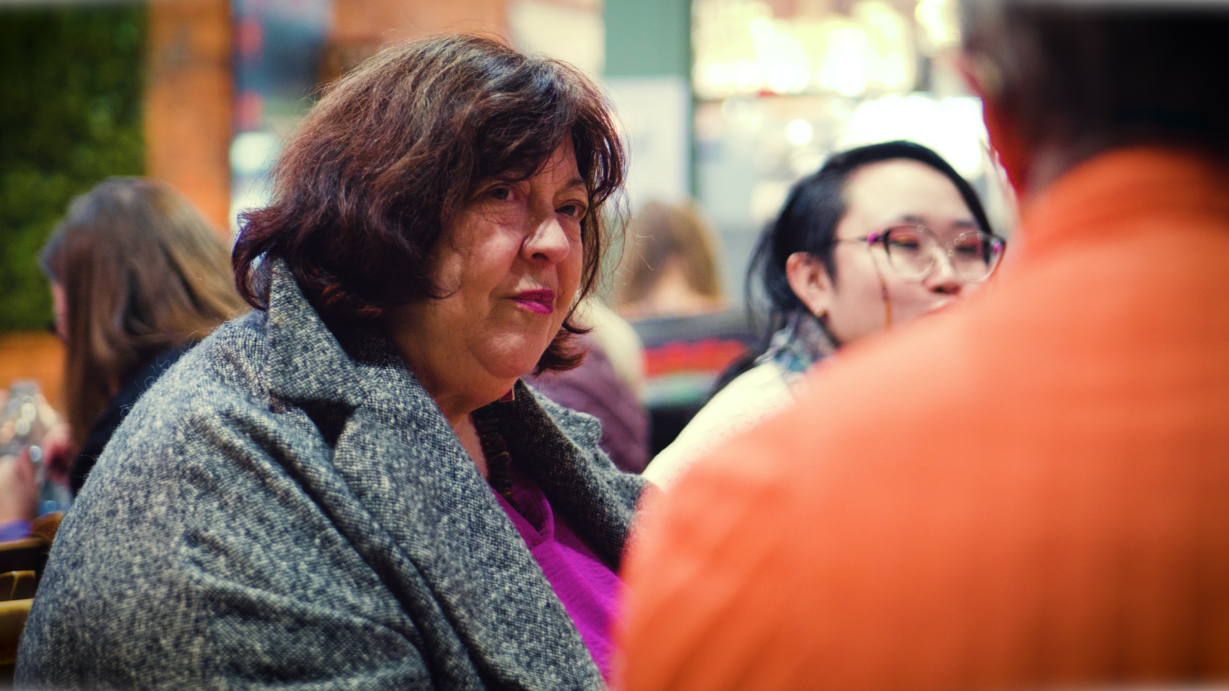 A middle-aged woman with dark brown, wavy hair and wearing a gray coat is looking to her left at a social gathering. There are other people in the background, including a woman with dark hair and glasses and a person wearing an orange top, seated in 
