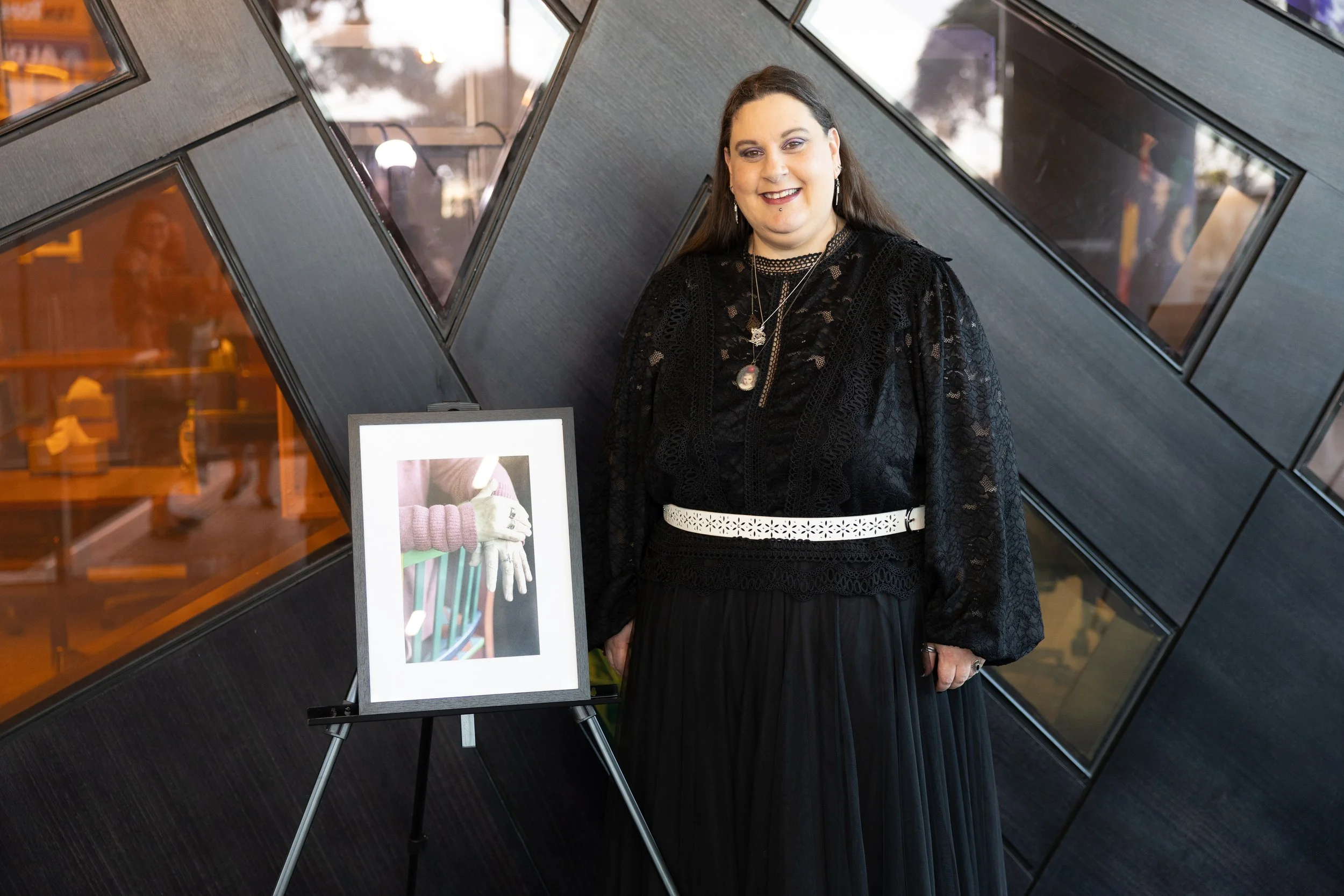 A woman dressed in black standing next to a framed photo of hands wearing gloves, against a modern black wall with angular windows.