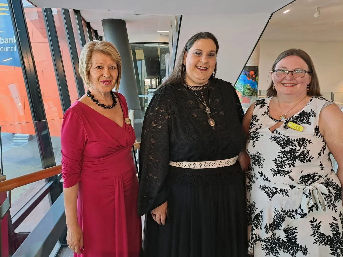 Three women standing indoors near large windows, posing for a photo at an event. The woman on the left is wearing a red dress and a necklace, the woman in the middle is wearing a black lace dress, and the woman on the right is wearing a white dress w