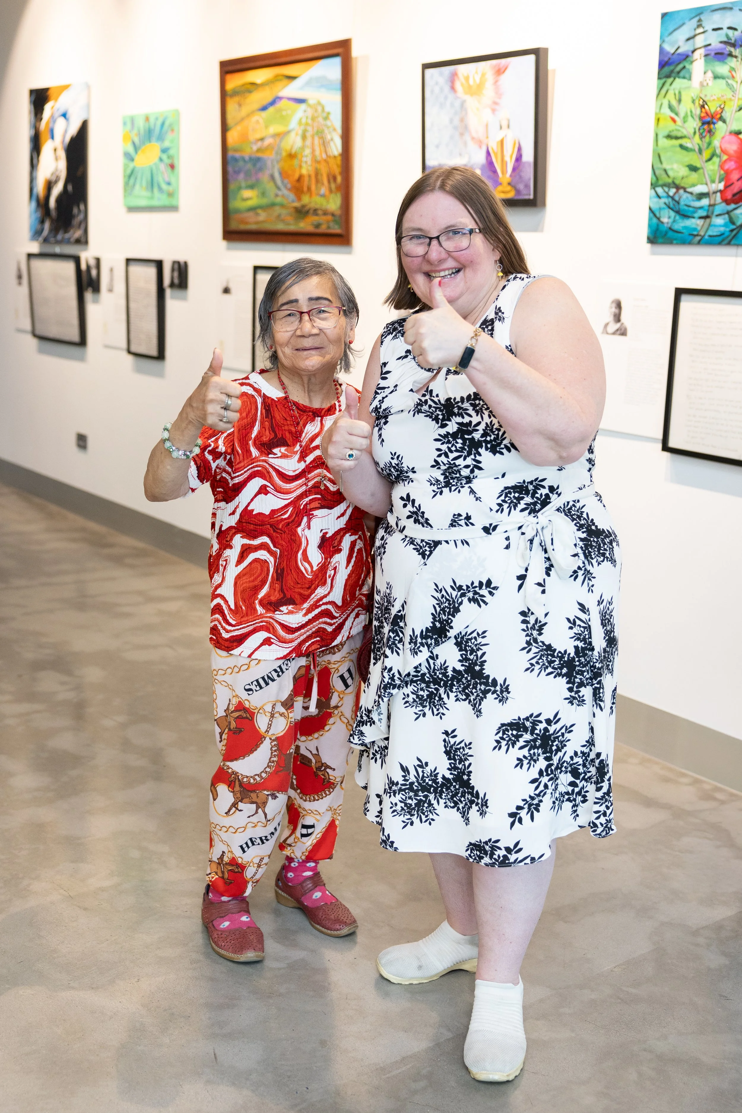 Two women standing in an art gallery, smiling and giving thumbs up. One woman is wearing a red and white patterned top with matching pants, and the other is dressed in a white dress with black floral patterns.
