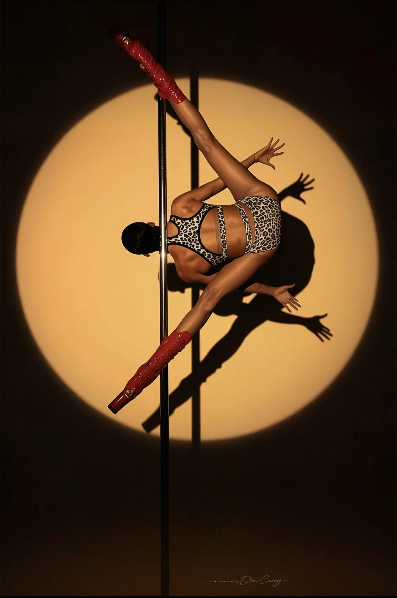 A pole dancer performing on stage, illuminated by a spotlight creating a circular shadow behind her. She wears a leopard print outfit and red thigh-high boots.