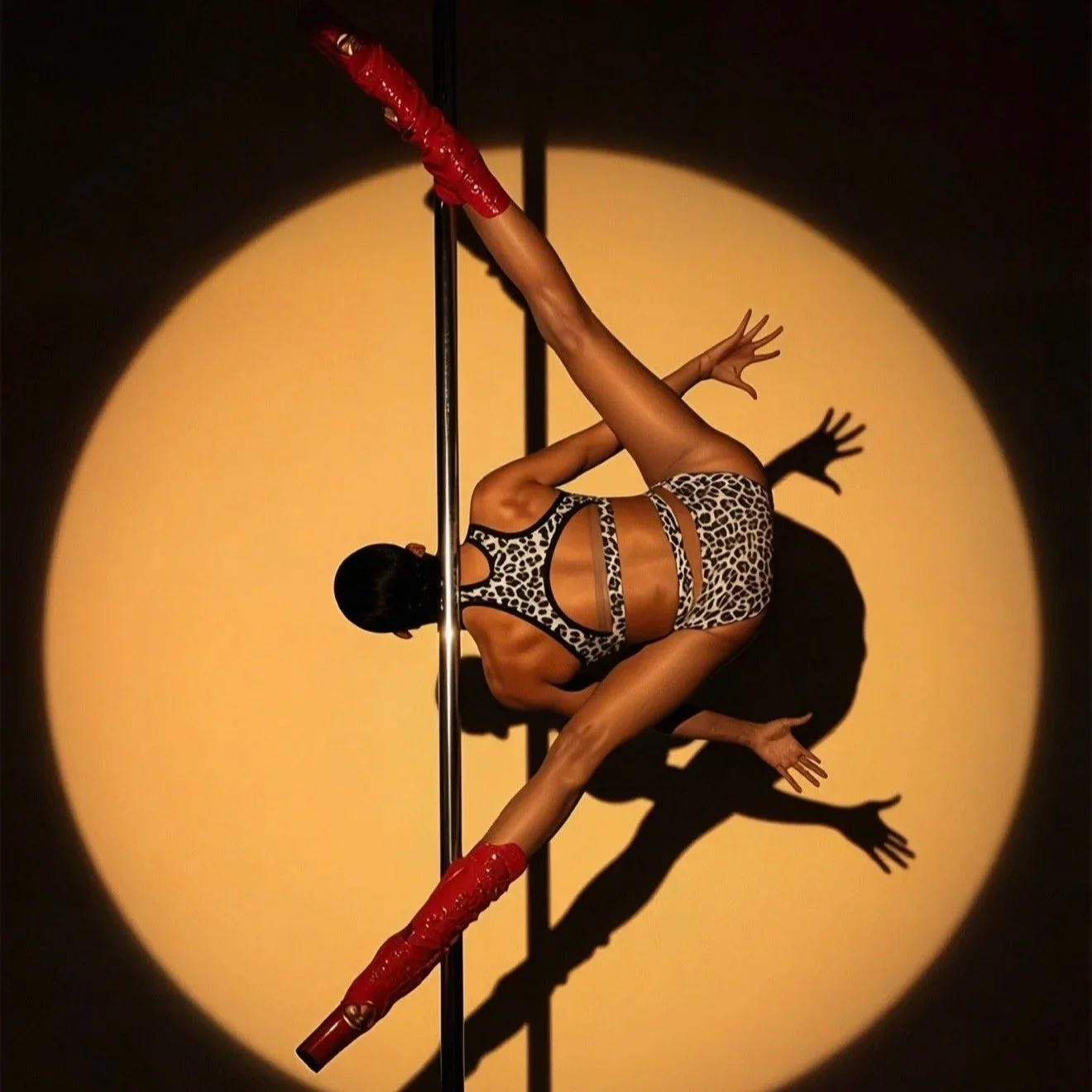 A female pole dancer performing on a stage, dressed in a leopard print costume with red gloves and boots, against a yellow spotlight with her shadow cast on the wall.