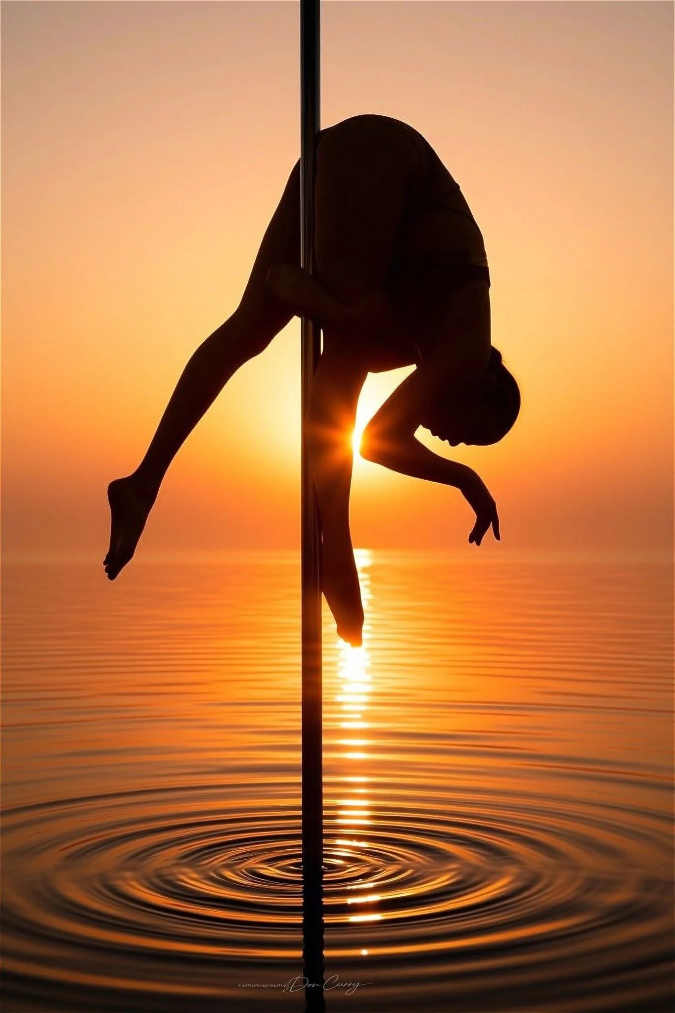 Silhouette of a person performing a pole dance move during sunset over a body of water.