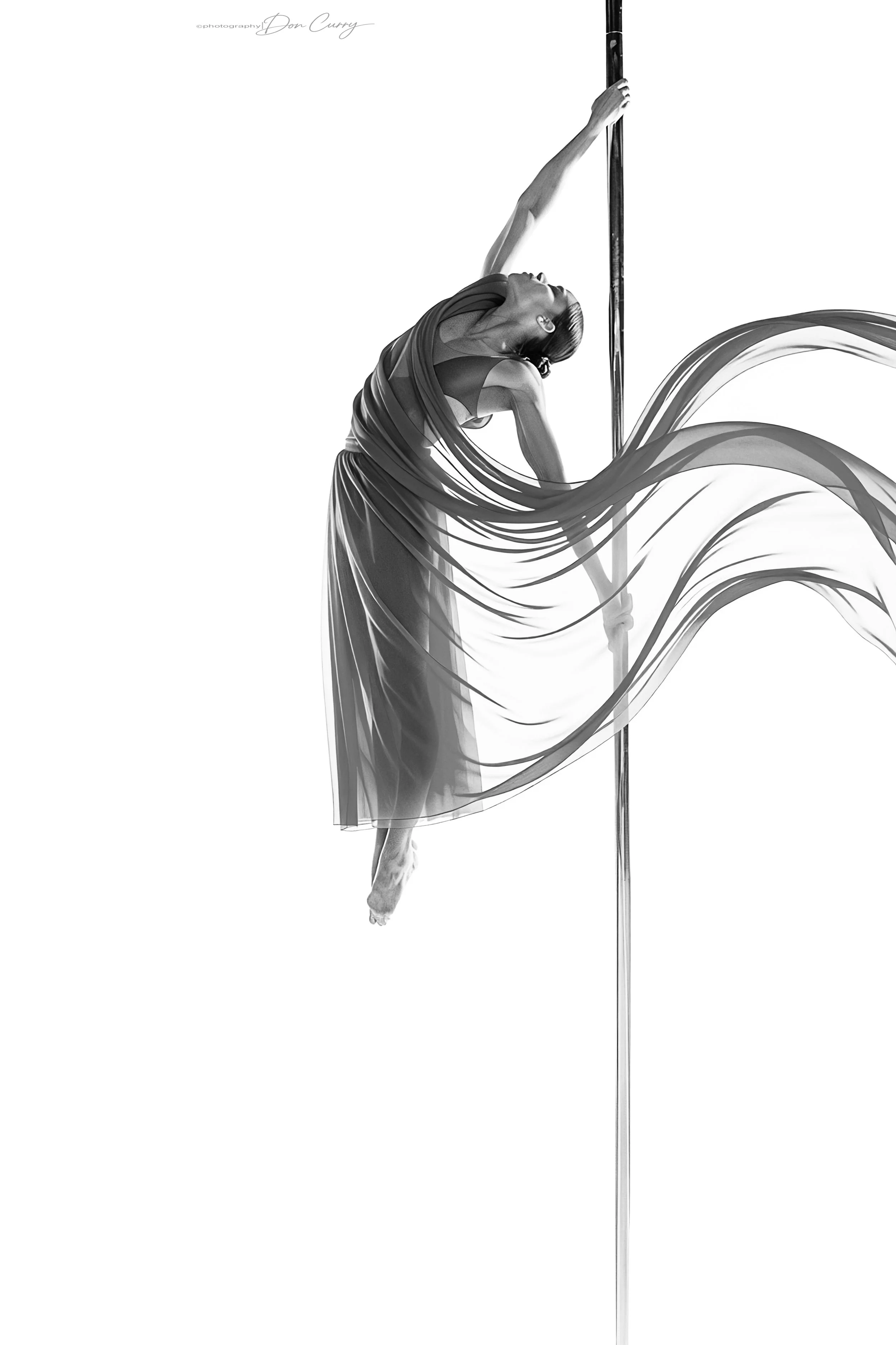 Black and white photo of a woman pole dancing, wearing a flowing skirt and a sports bra, mid-air with her head tilted back.