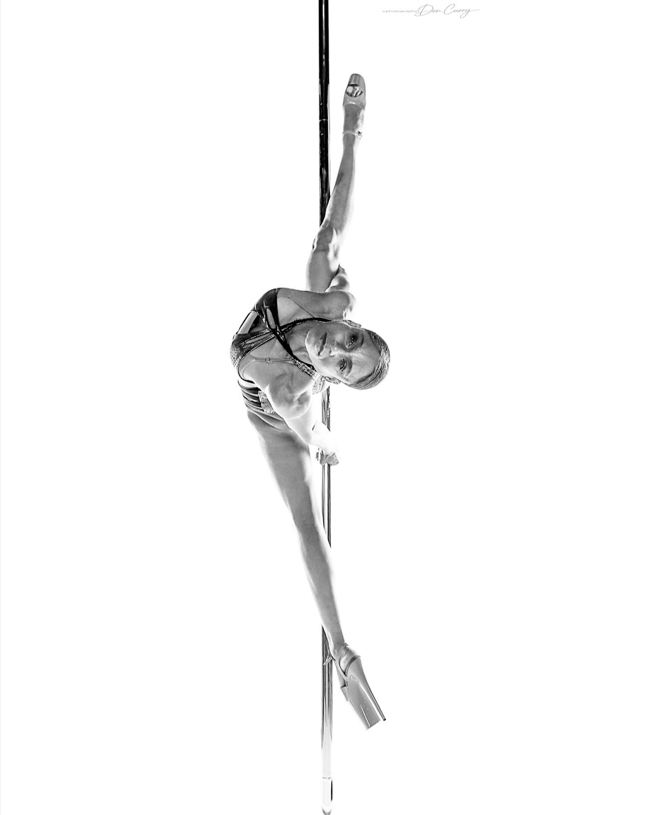 A female pole dancer performing an acrobatic move on a vertical pole in black and white.