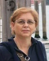 Mulher com cabelo castanho claro, óculos e expressão neutra, ao ar livre próximo a uma grade de ferro.