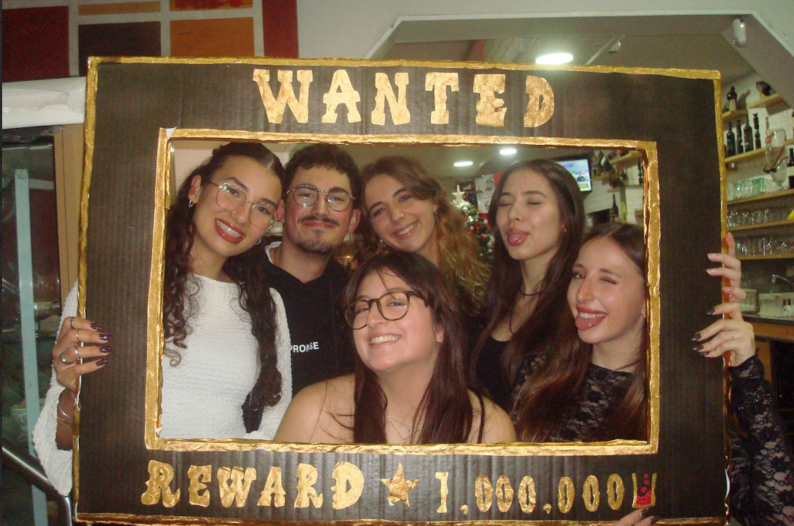 Grupo de seis pessoas sorrindo e brincando com uma moldura de quadro falsa com a inscrição 'WANTED REWARD 1,000,000' em um ambiente de bar ou restaurante decorado com tema natalino.
