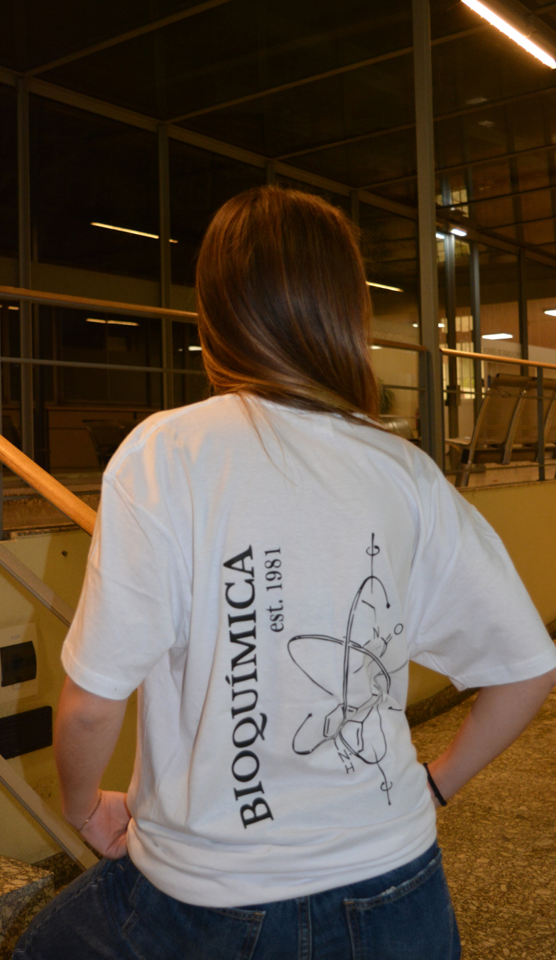 Pessoa com camiseta branca de costas, com o texto 'Bioquímica est. 1981' e um desenho de molécula pequena, na parte de trás, em um ambiente interno moderno com janelas de vidro.