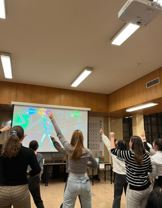 Grupo de pessoas jovens participando de um jogo de dança em uma sala de aula ou espaço de lazer, assistindo a uma tela com uma personagem animada.