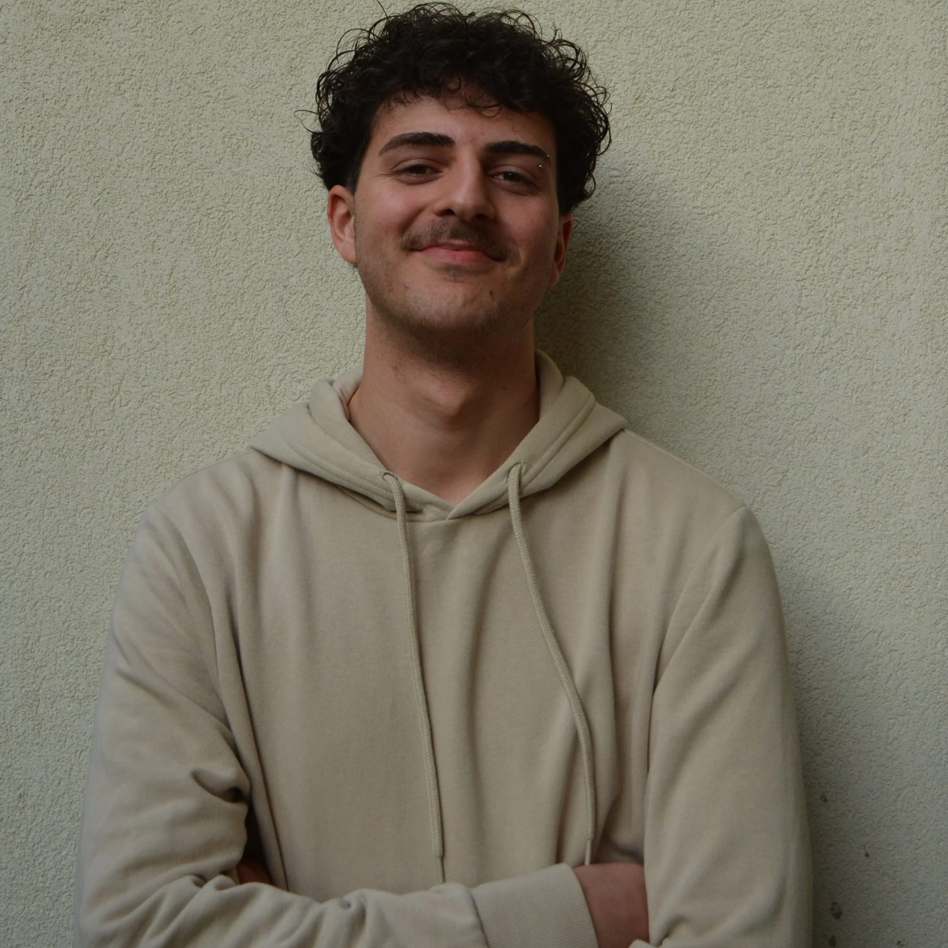 Jovem sorridente com cabelo cacheado, usando moletom bege, de braços cruzados, posando contra uma parede de cor clara.