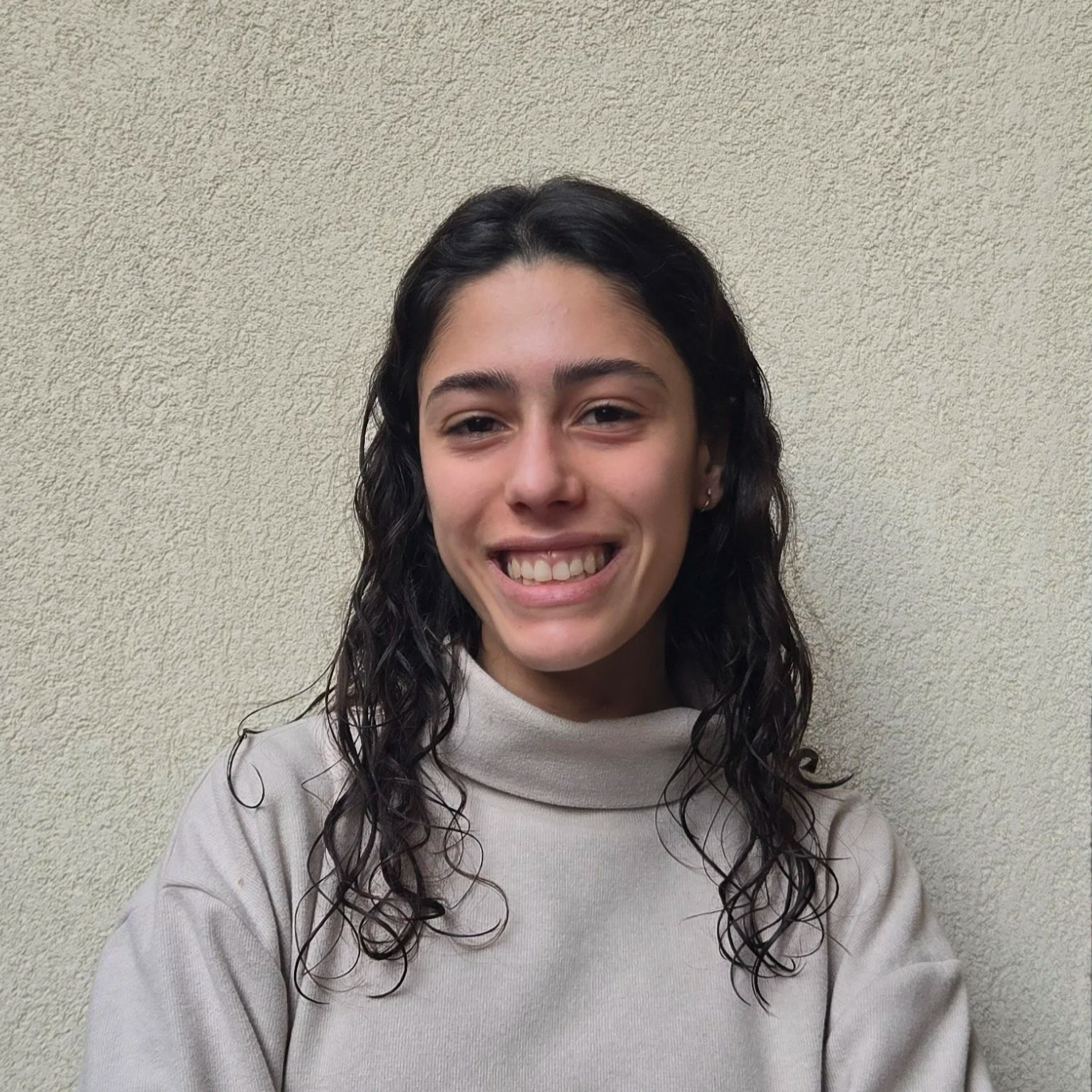 Jovem de cabelo cacheado escuro, sorridente, usando suéter bege, posando contra parede de textura leve e cor clara.