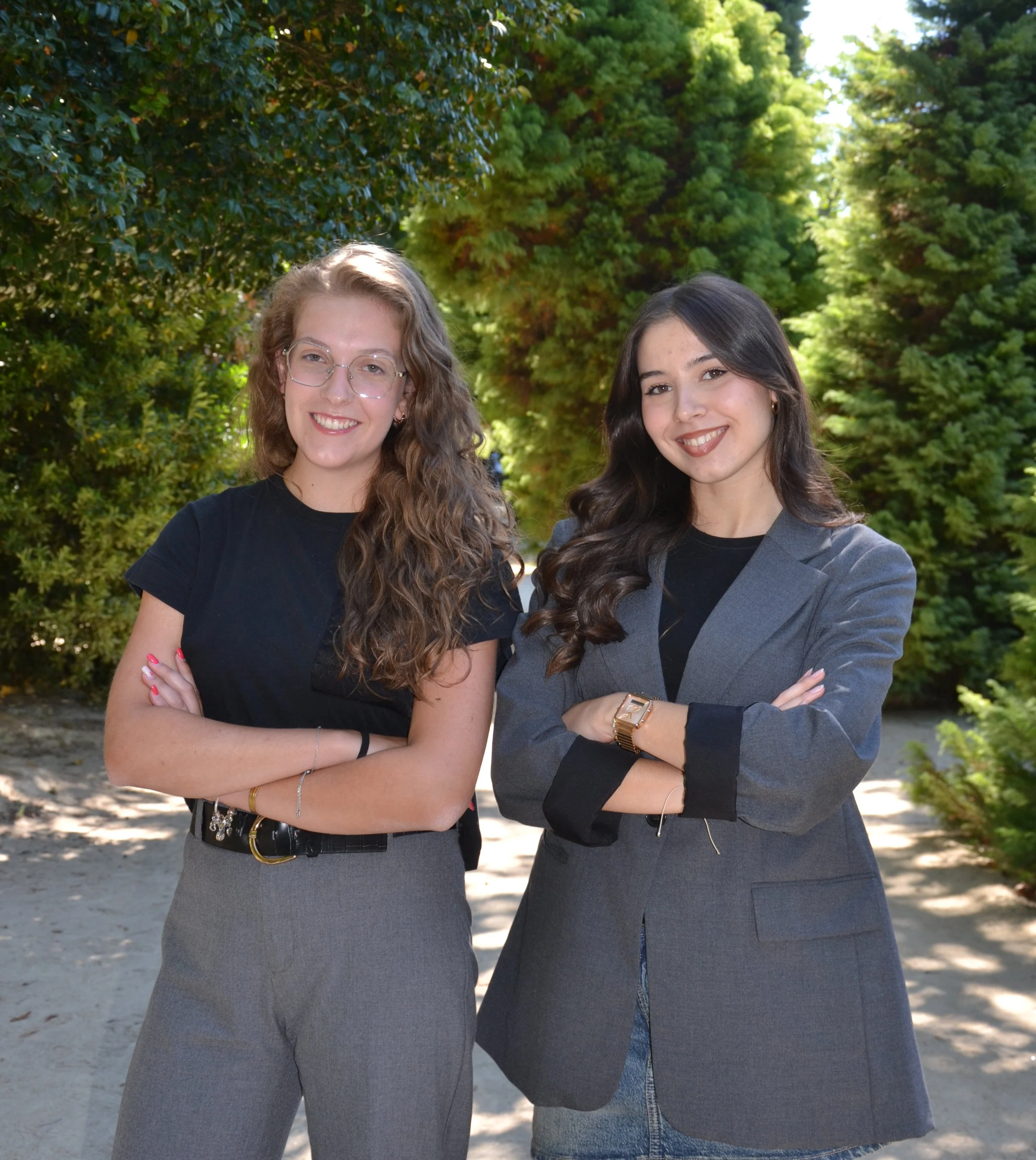 Duas mulheres jovens sorrindo, uma com cabelo encaracolado, usando óculos e uma blusa preta, a outra com cabelo liso, usando terno cinza e jaqueta preta, com fundo de árvores verdes.