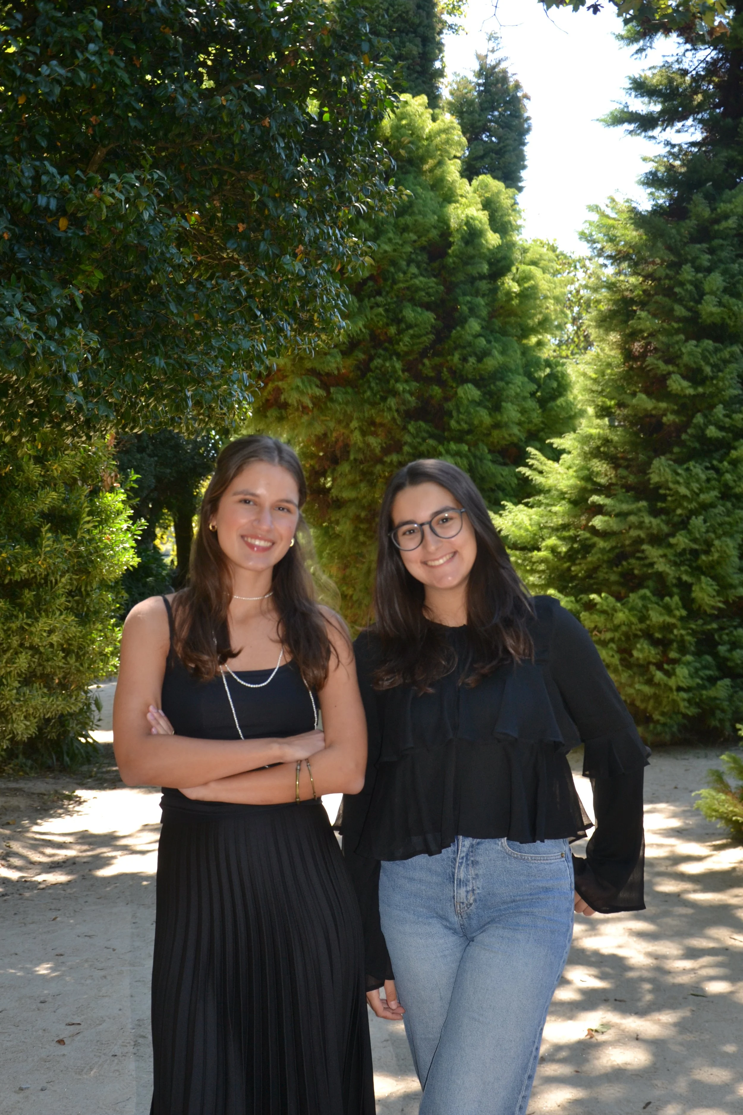 Duas jovens mulheres sorridentes posando juntas ao ar livre, com árvores verdes ao fundo, em um dia ensolarado.