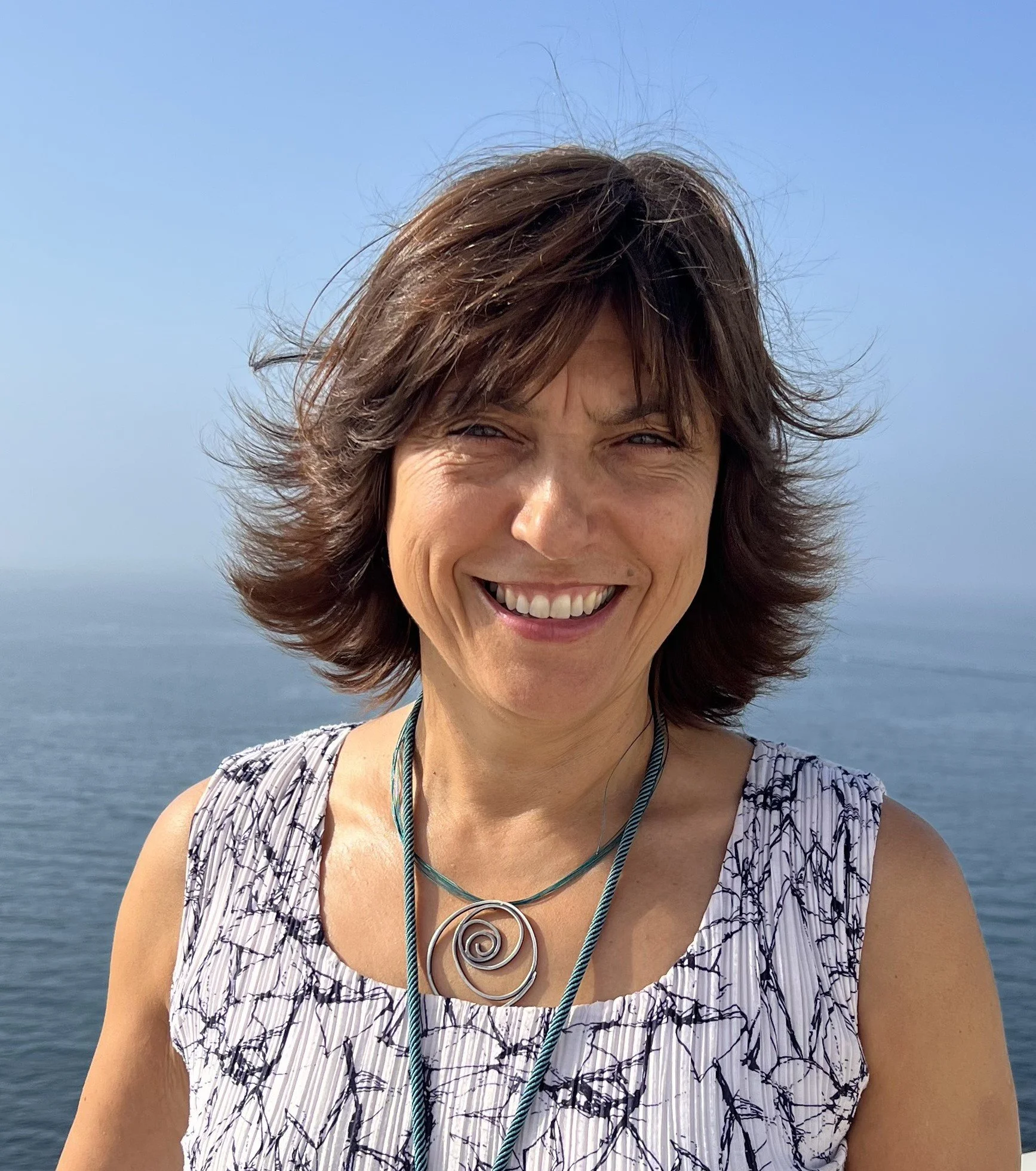 Mulher sorrindo ao ar livre com mar ao fundo, usando blusa de verão e colar com fecho de espiral.