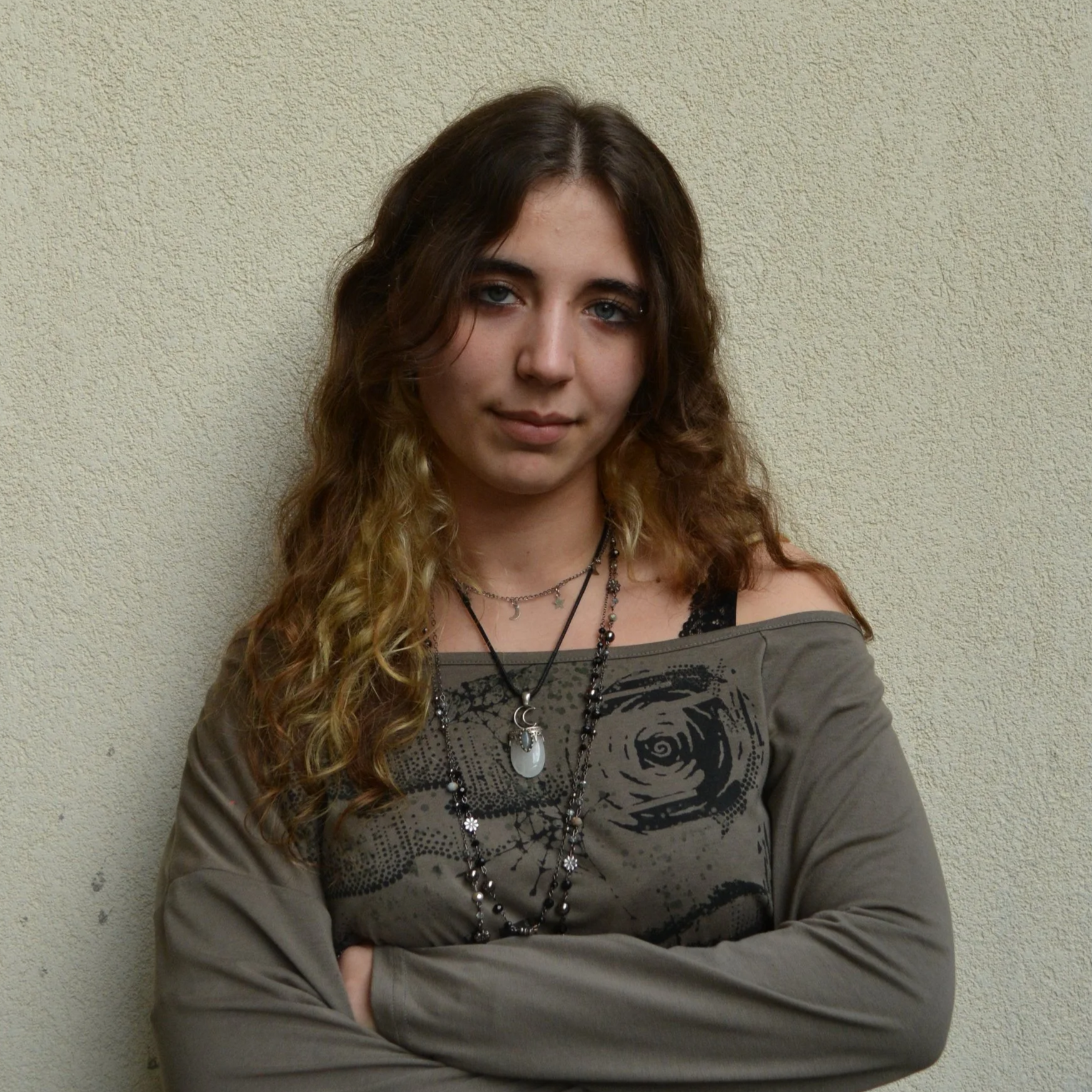 Jovem de cabelos castanhos ondulados e olhos azuis, vestindo camisa cinza com estampa, várias colares e de braços cruzados posando em frente a uma parede beige.