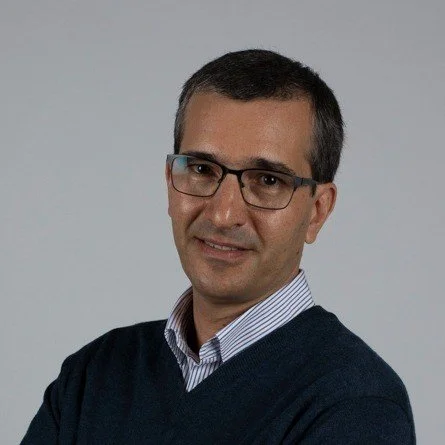 Homem com óculos, cabelo curto e escuro, vestindo camisa de botões e suéter escuro, sorrindo com fundo cinza claro.
