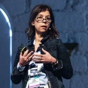 Mulher com óculos, cabelo escuro e vestido xadrez, segurando um tablet e falando em um palco com fundo de parede de pedra