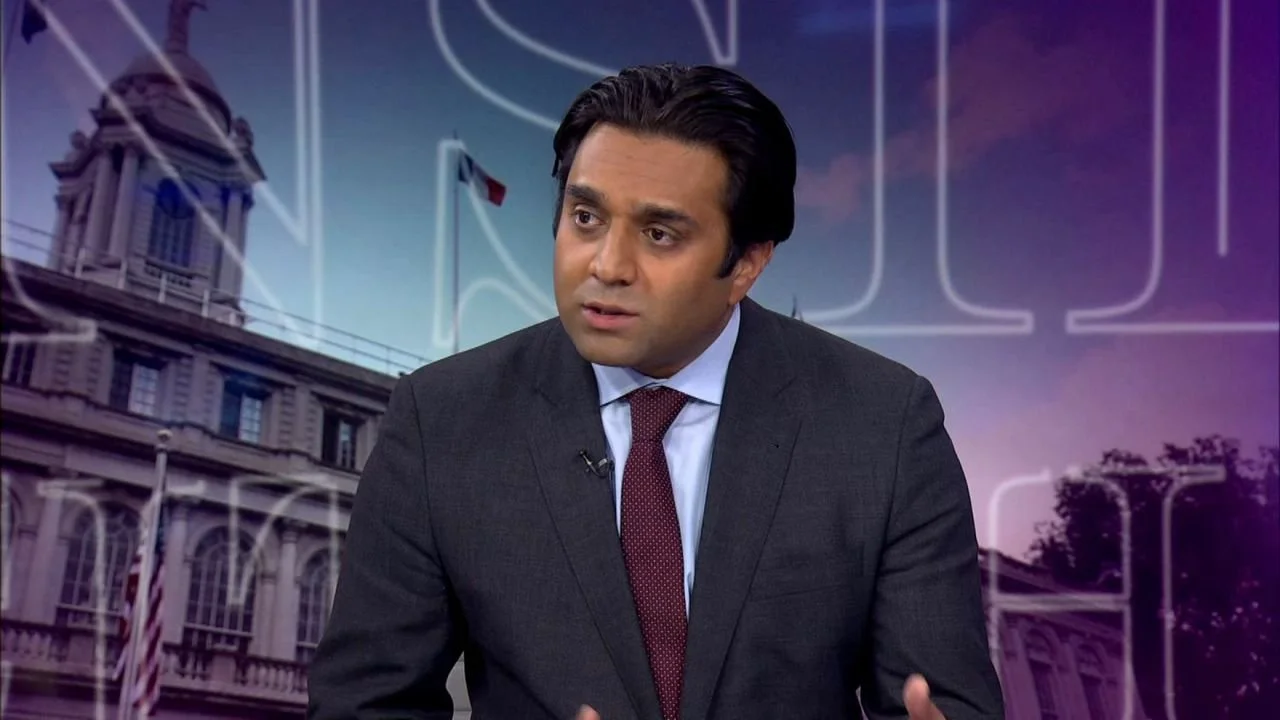 A man in a dark suit with a white shirt and patterned red tie, speaking during a television interview or talk show with a city hall building and French flags in the background.
