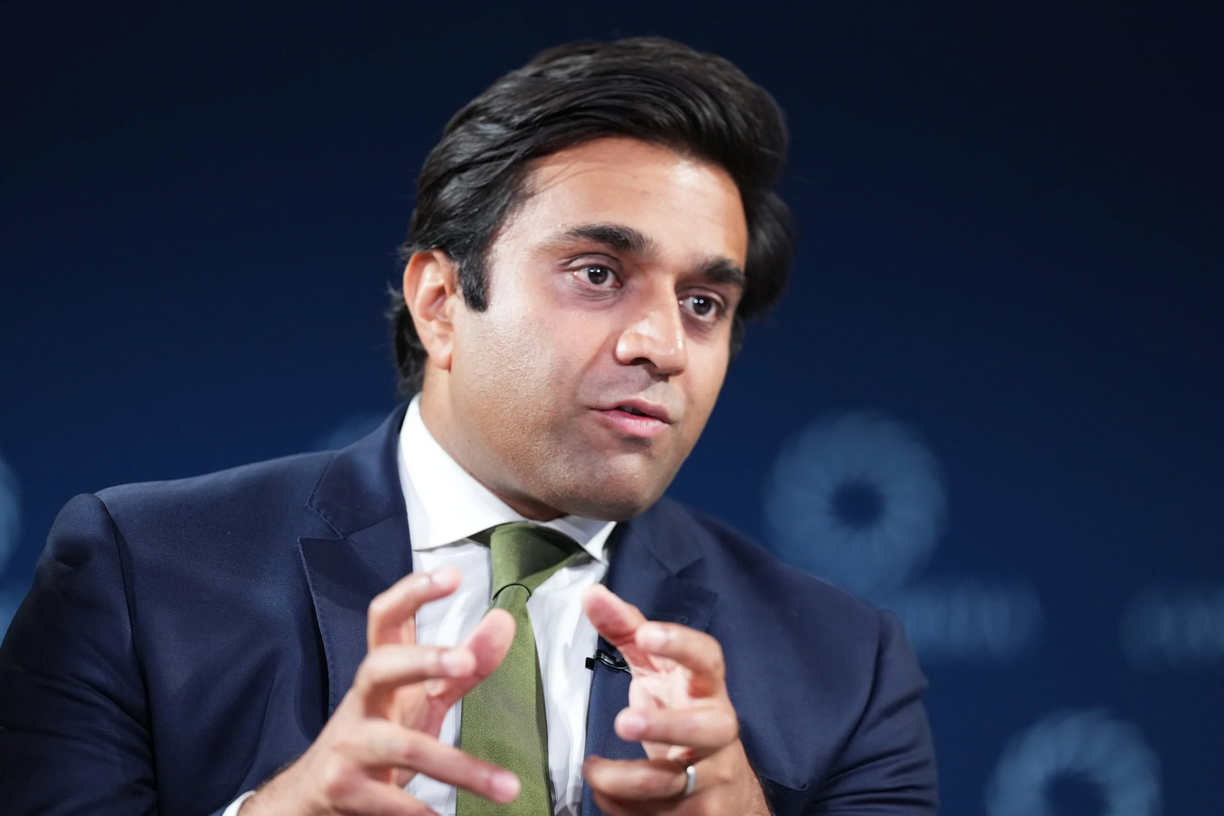 A man in a suit and tie speaking or gesturing with his hands against a dark background.