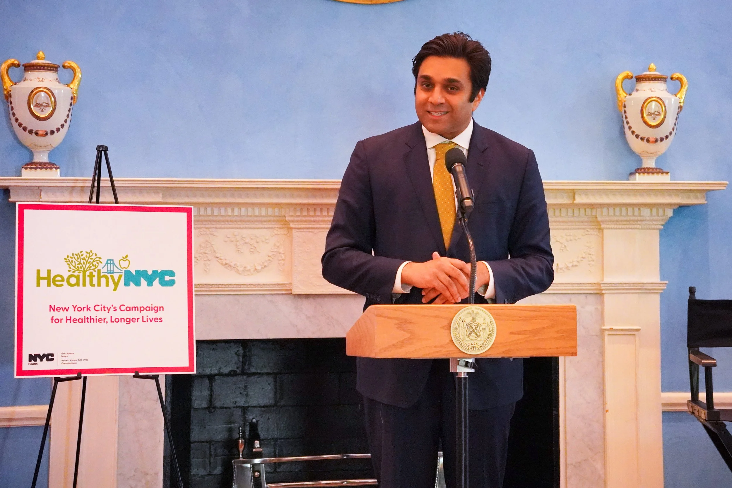 A man in a dark suit and yellow tie standing at a podium, speaking into a microphone, in a room with light blue walls and decorative vases on a mantel behind him, with a sign reading "Healthy NYC" and "New York City’s Campaign for Healthier, Longer Lives" in front of him.