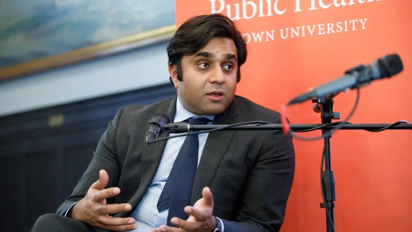Ashwin Vasan in a gray suit and white shirt speaking into a microphone at a panel or discussion. Behind him is an orange background with text partially visible pointing to a university.