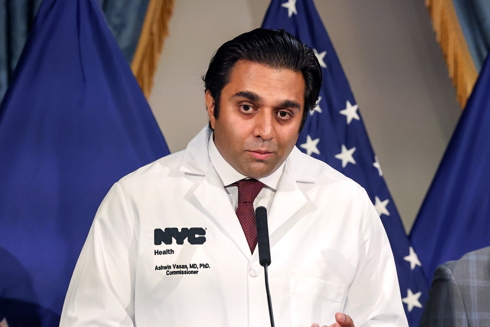 Ashwin Vasan in a white medical coat standing behind a microphone with an American flag in the background. The coat has text that reads 'NYC Health' and 'Ashwin Vasan, MD, PhD, Commissioner'.