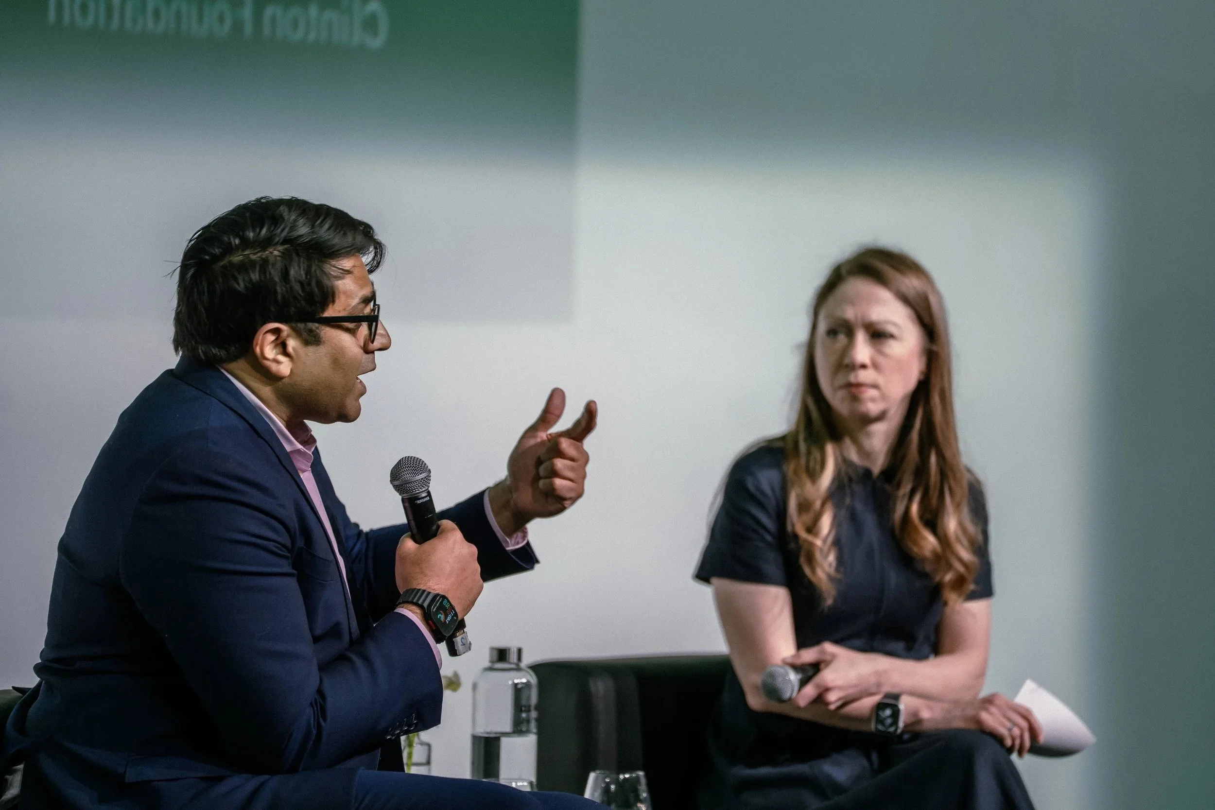 A man in a suit and glasses is speaking into a microphone, gesturing with his right hand, while a woman with long reddish hair and a dark blazer listens and looks at him. They are seated during a discussion or presentation, with microphones and water bottles visible.