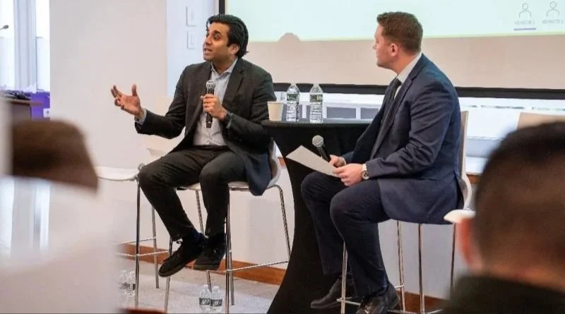 Ashwin Vasan to the left and another man to the right, both in suits seated on stage during a panel discussion or interview, one speaking while holding a microphone, and the other listening with a notebook, in a professional setting.