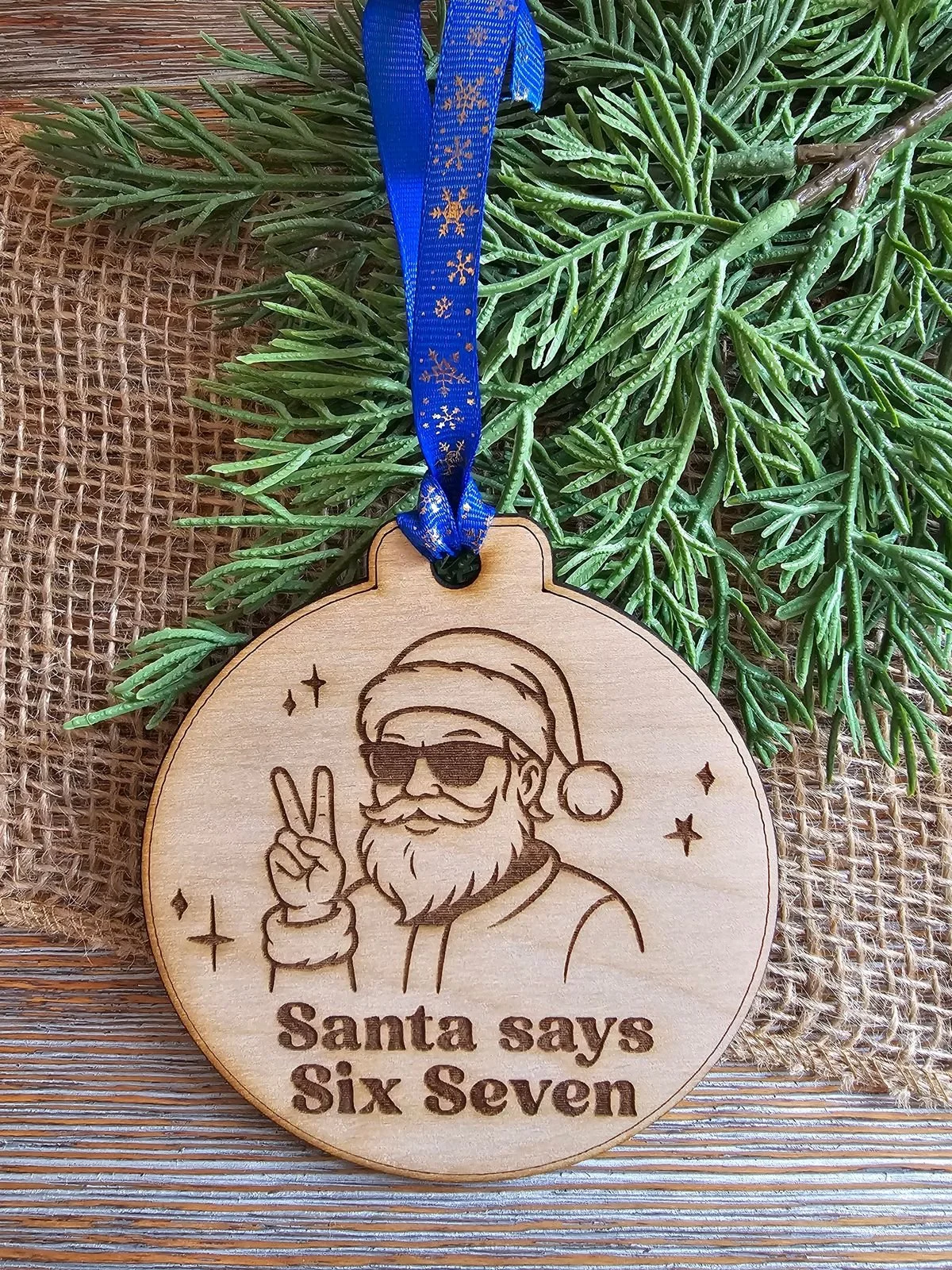 A wooden Christmas ornament with a drawing of Santa wearing sunglasses and holding up a peace sign, engraved with the words "Santa says Six Seven." It hangs from a blue ribbon decorated with gold snowflakes, placed on green foliage and burlap.