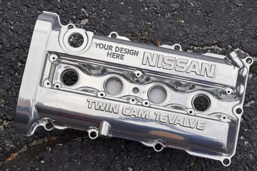 Shiny silver Nissan engine valve cover with customizable text, placed on asphalt. Fixed text includes 'NISSAN', 'TWIN CAM 16 VALVE', and 'YOUR DESIGN HERE'.