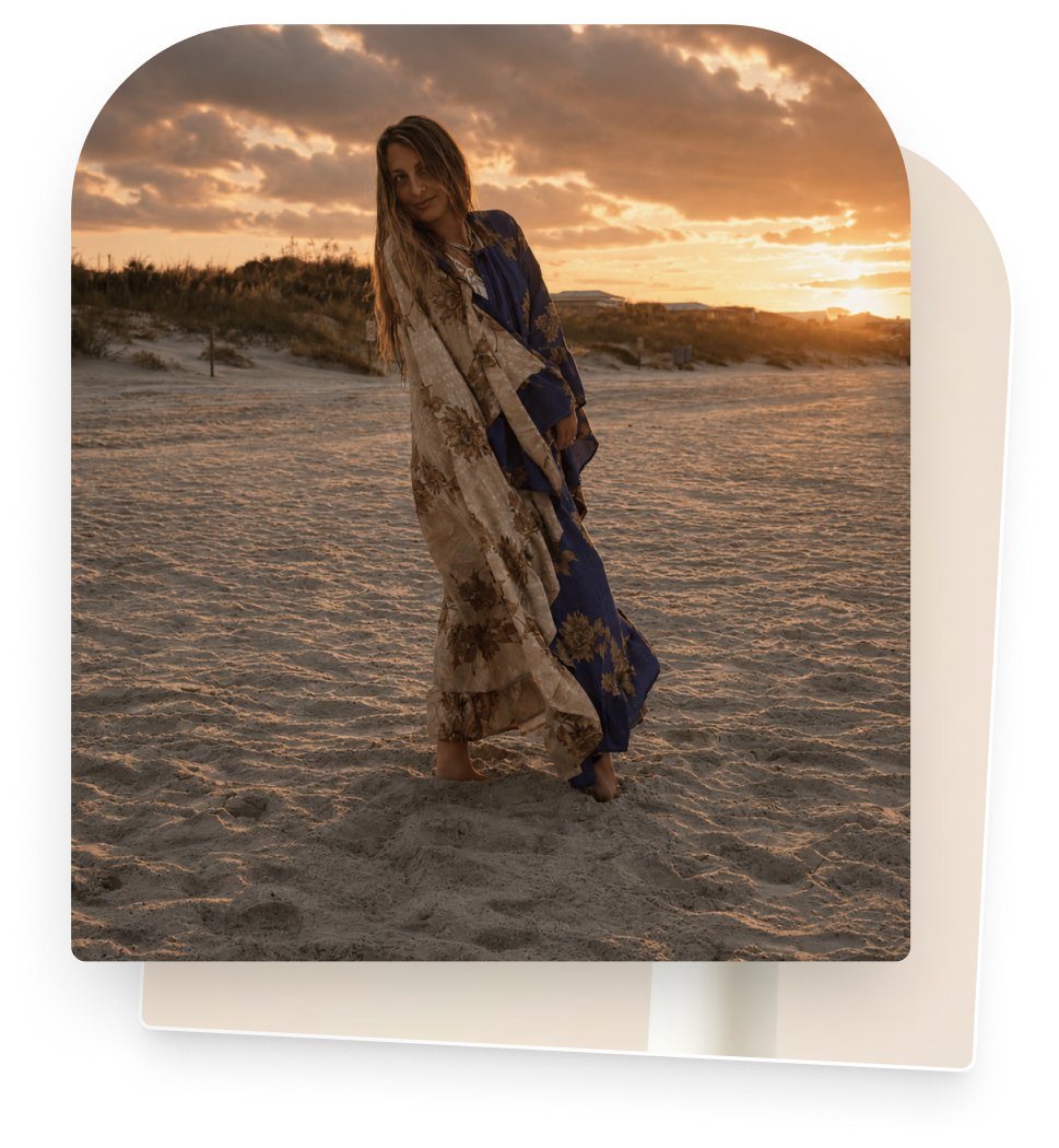 Woman standing on a sandy beach during sunset with her hair and a patterned dress flowing in the breeze.