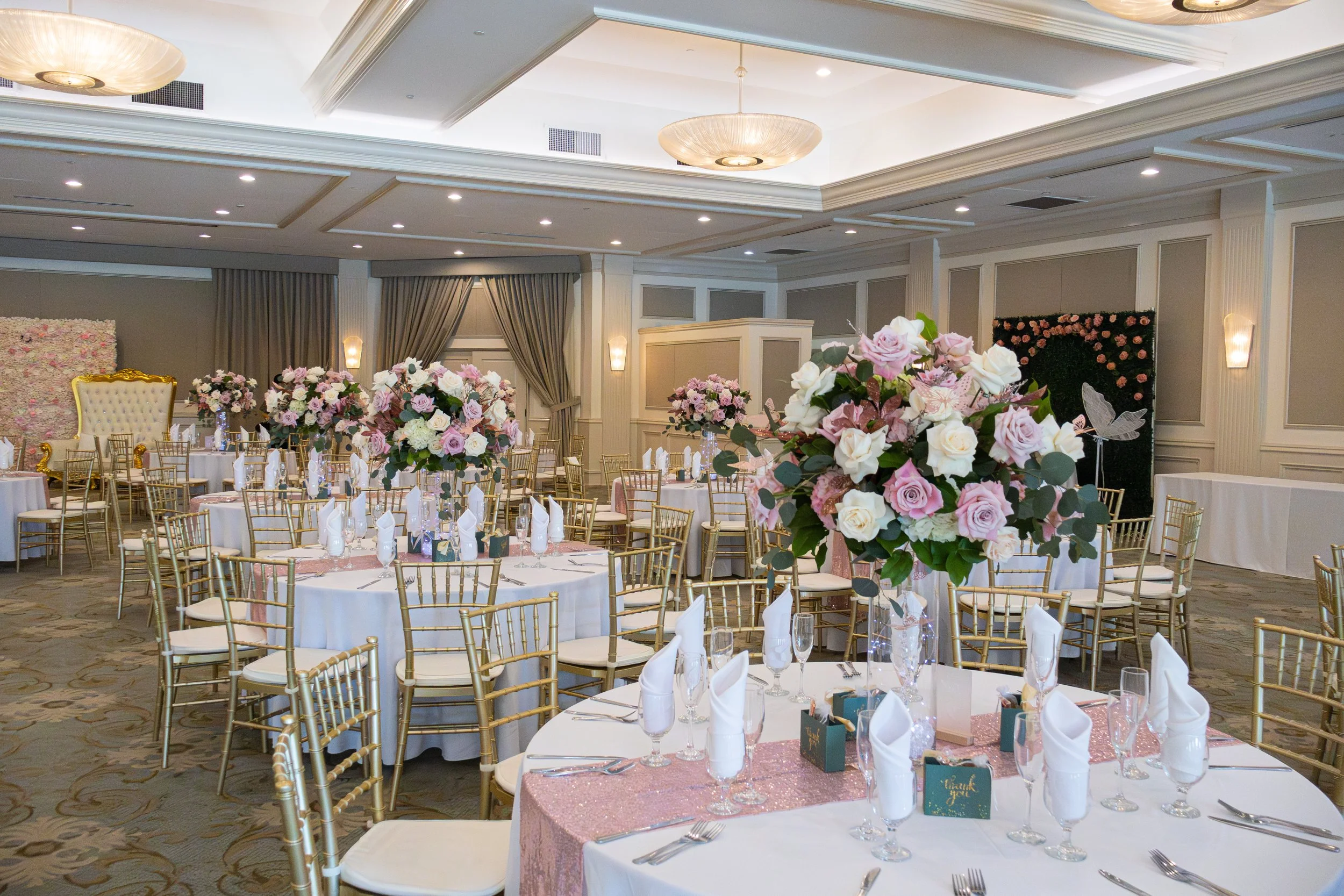 Elegant banquet hall decorated for a wedding reception with round tables, white tablecloths, pink and white floral centerpieces, gold chairs, and pink sequined table runners.
