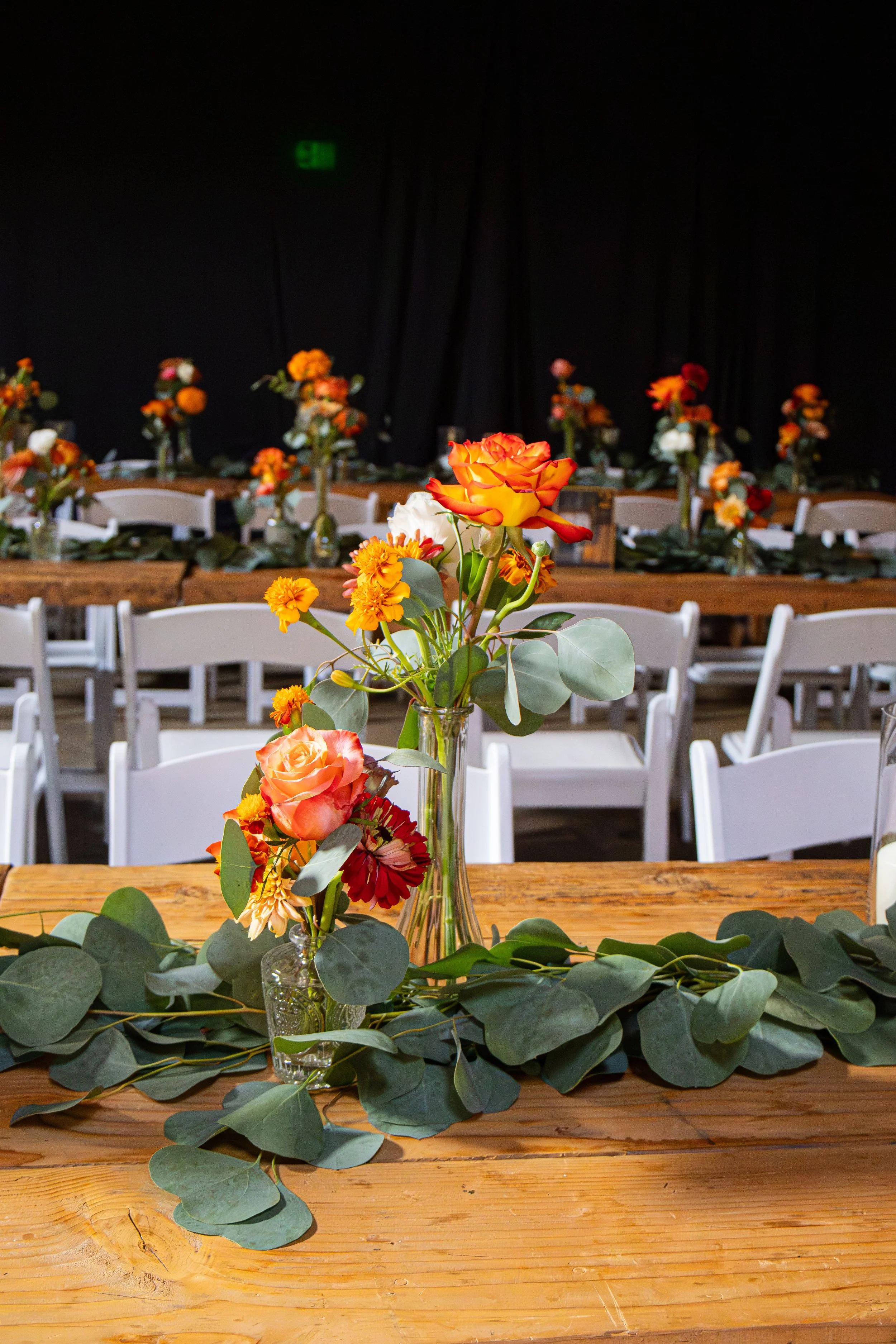Centerpieces with orange, peach, and yellow flowers on a wooden table, surrounded by chairs, with more floral arrangements in the background, indicating a decorated event or reception hall.