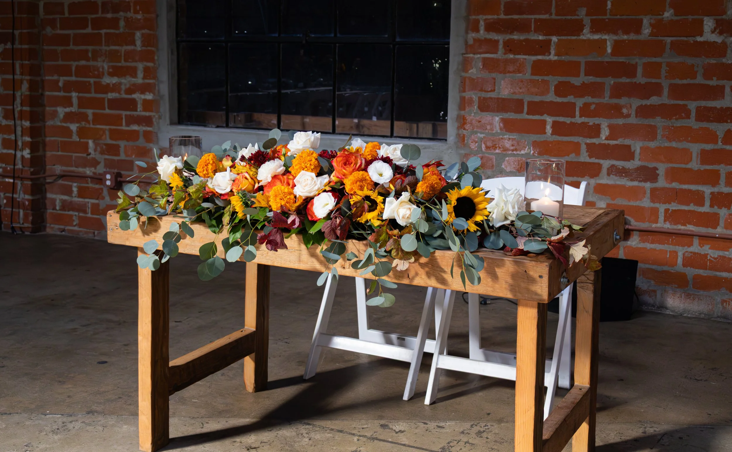 A wooden table decorated with an arrangement of flowers and foliage, and lit candles, in an industrial-style room with exposed brick walls and a window.