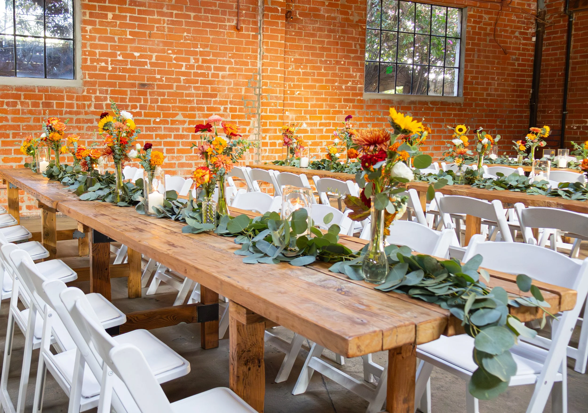 Long wooden table decorated with colorful flower arrangements, candles, and greenery, set up for a wedding or event in a rustic brick-walled venue with large windows.