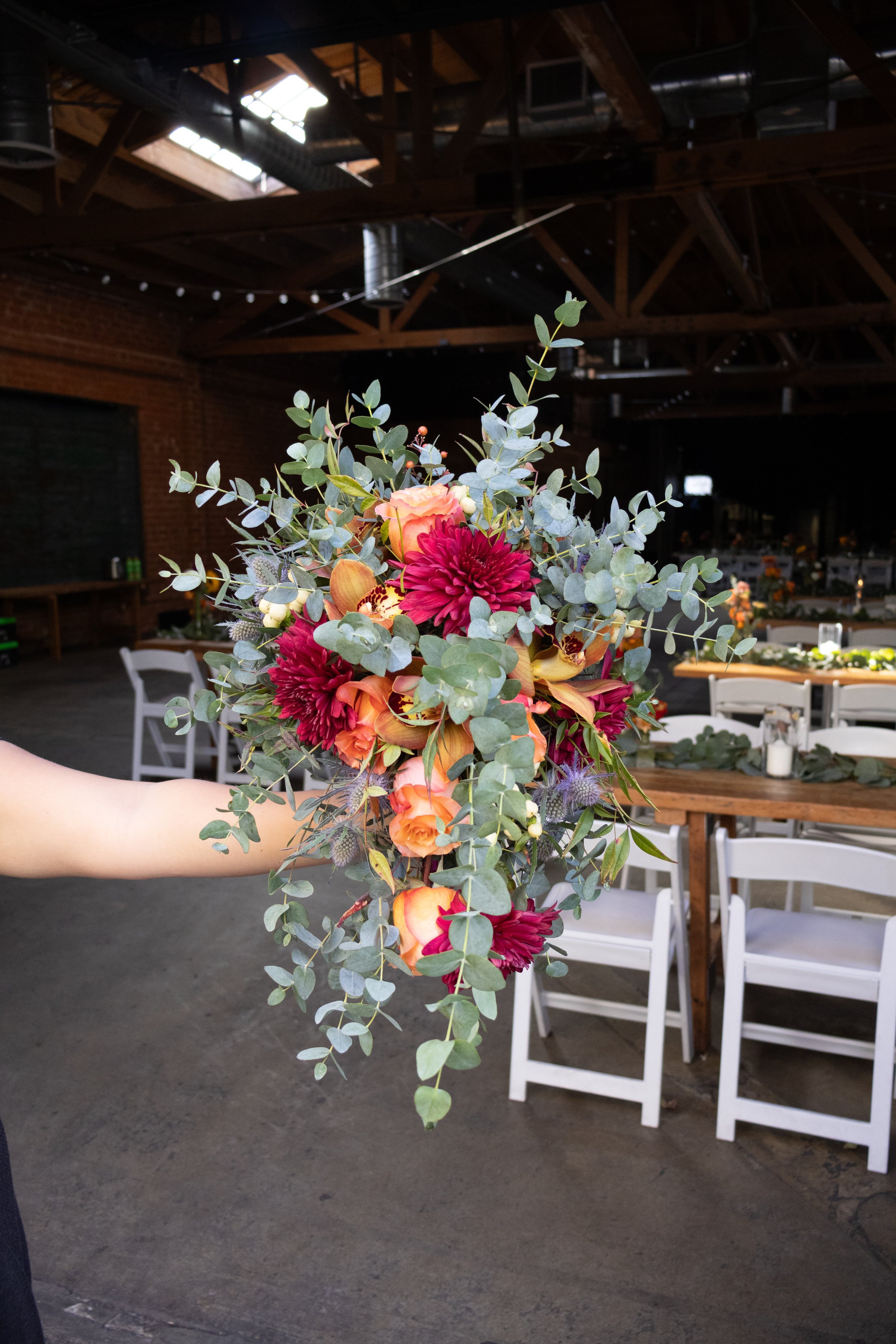 A person holding a cascading bouquet of orange, pink, and purple flowers with greenery inside a rustic venue with high wooden ceilings and white chairs.