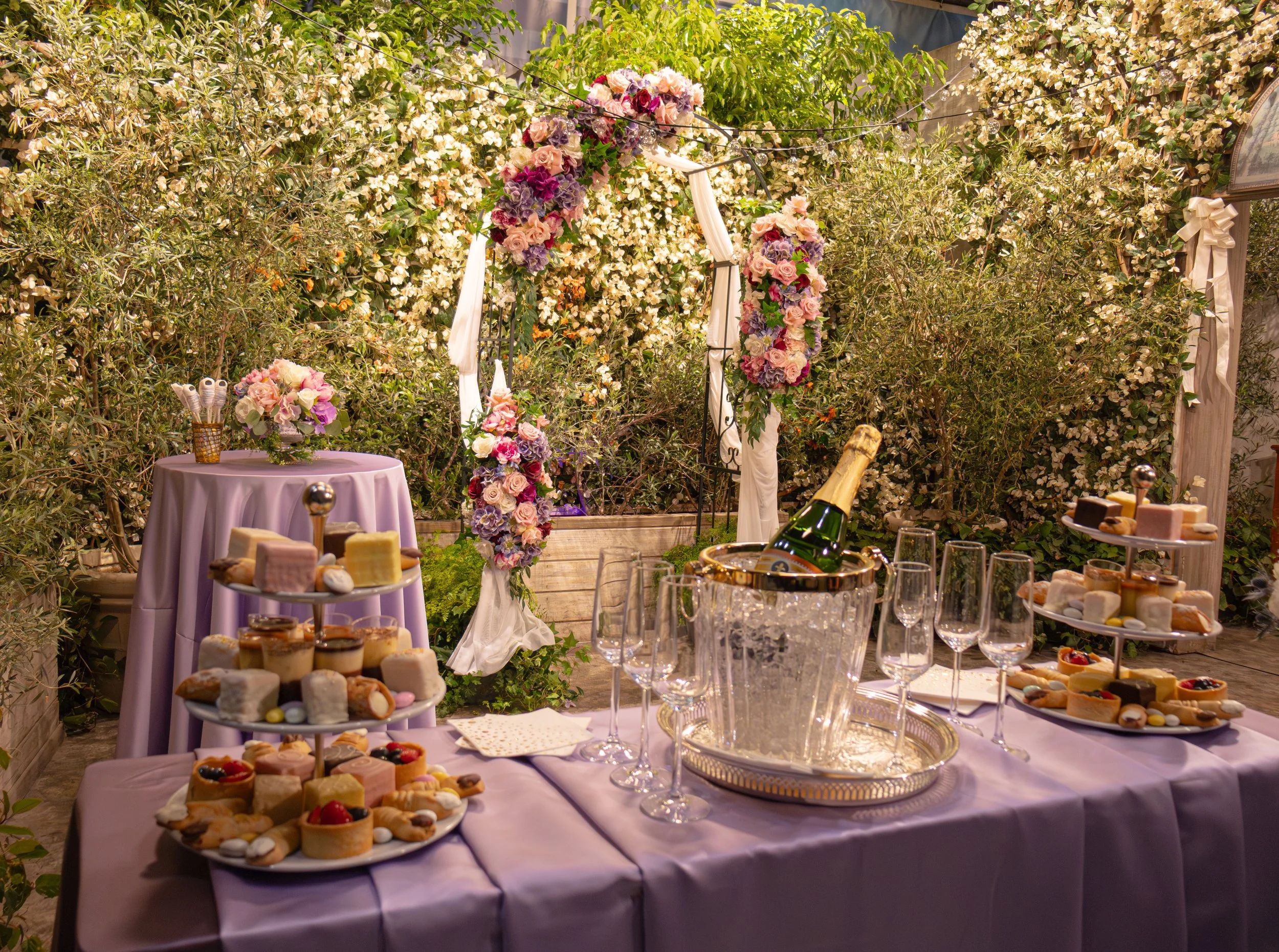 Decorative outdoor celebration setup with a table covered in lavender cloth featuring tiered trays of desserts, a champagne bottle in an ice bucket, empty glasses, and floral arrangements, with an arch decorated with flowers and greenery in the backg